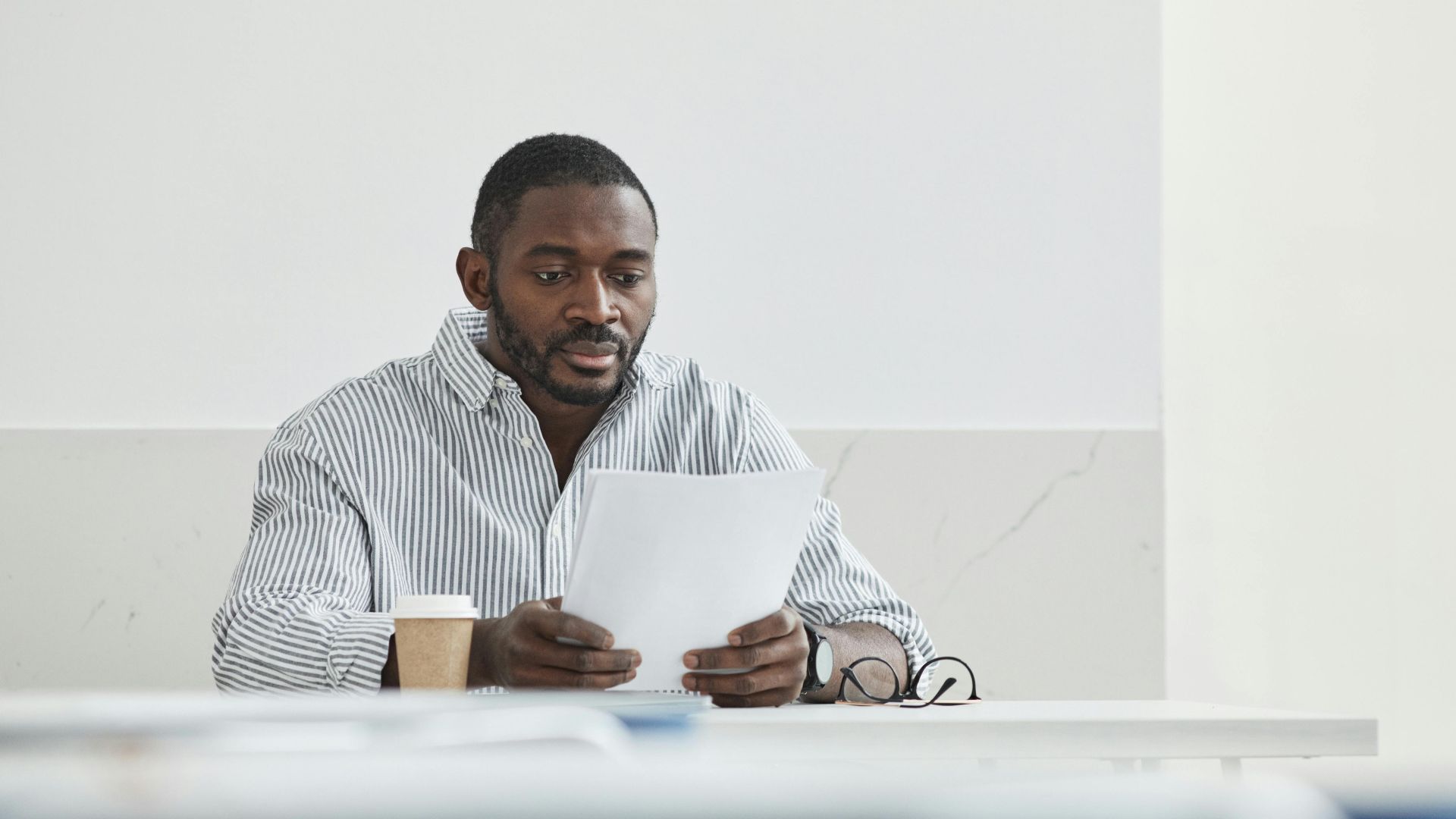African American man sitting indoors, reading papers with a coffee cup nearby.