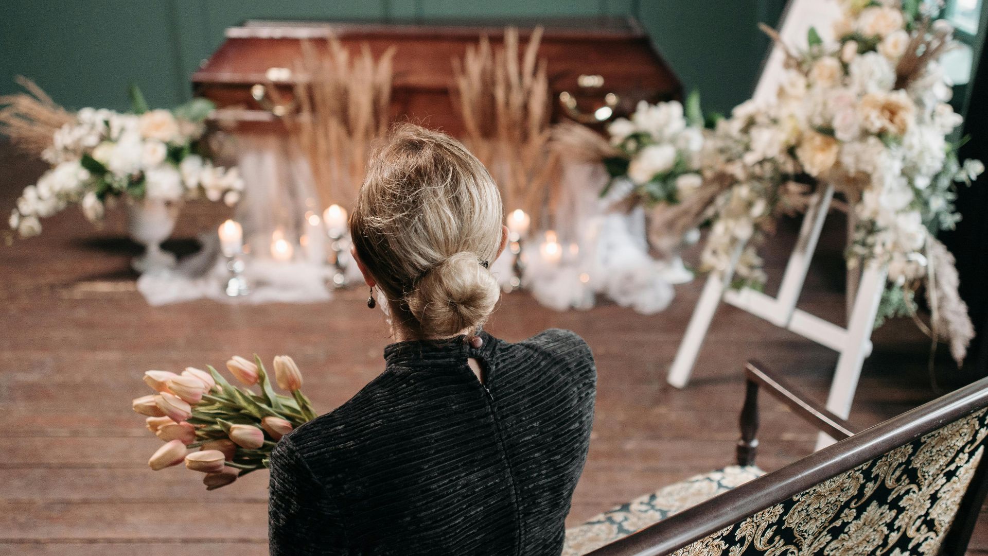 A woman mourns at an indoor funeral ceremony with floral decorations and a coffin.