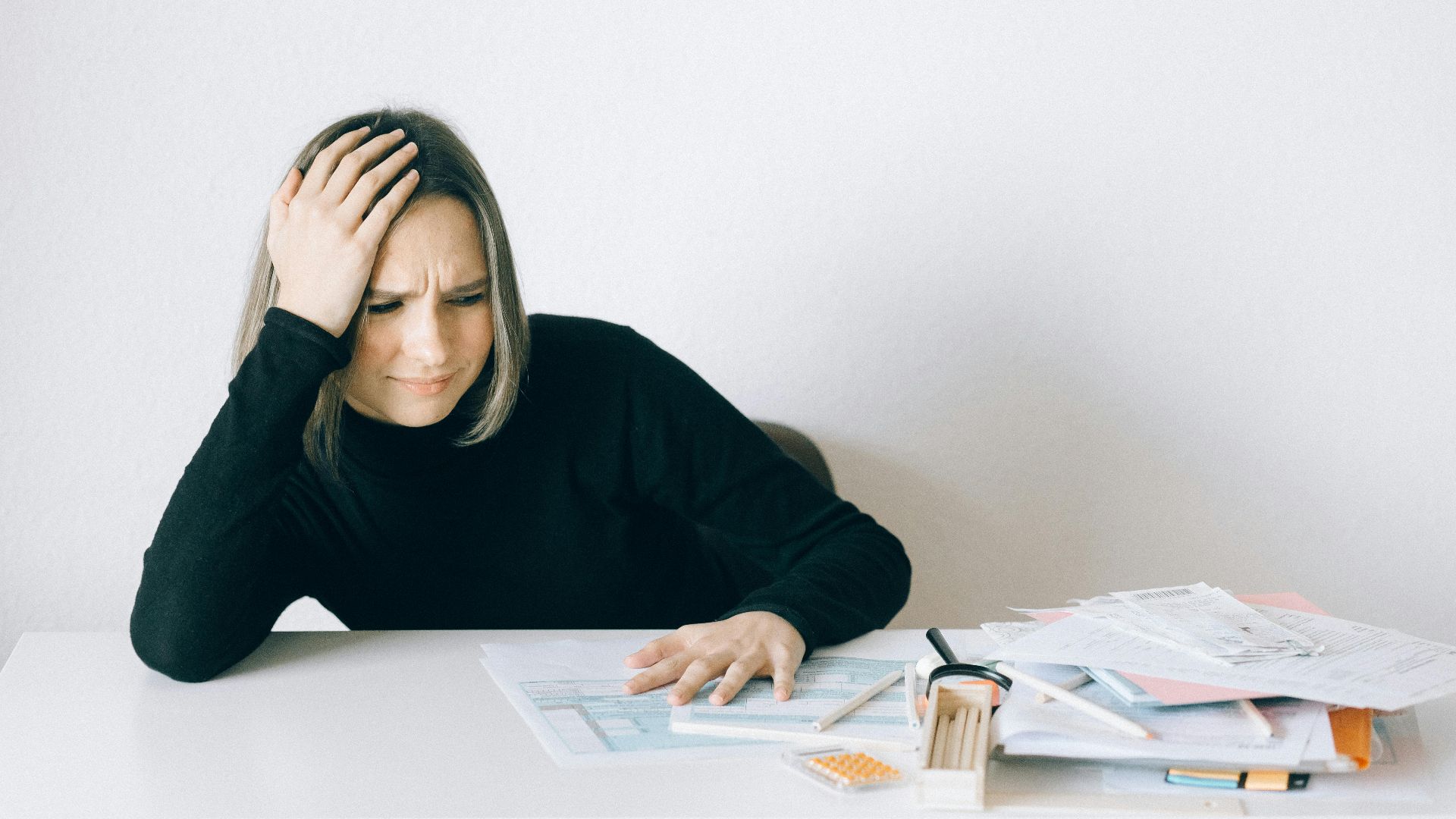 Woman in black sweater stressed with financial paperwork, overwhelmed at white table.
