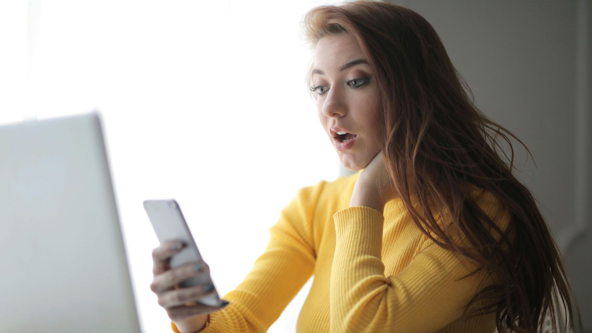 Young woman in yellow sweater looks surprised at phone while working on a laptop indoors.