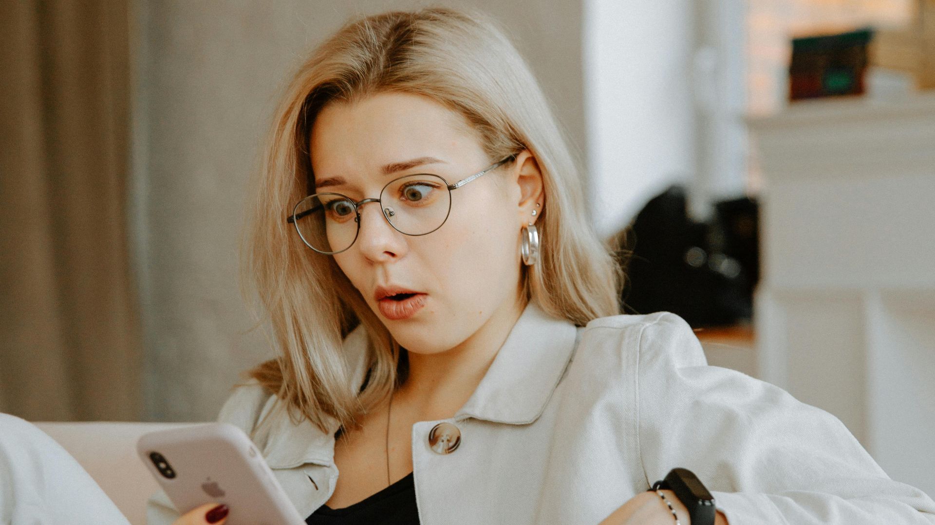 Surprised woman with glasses reacts to news on her phone while relaxing indoors.