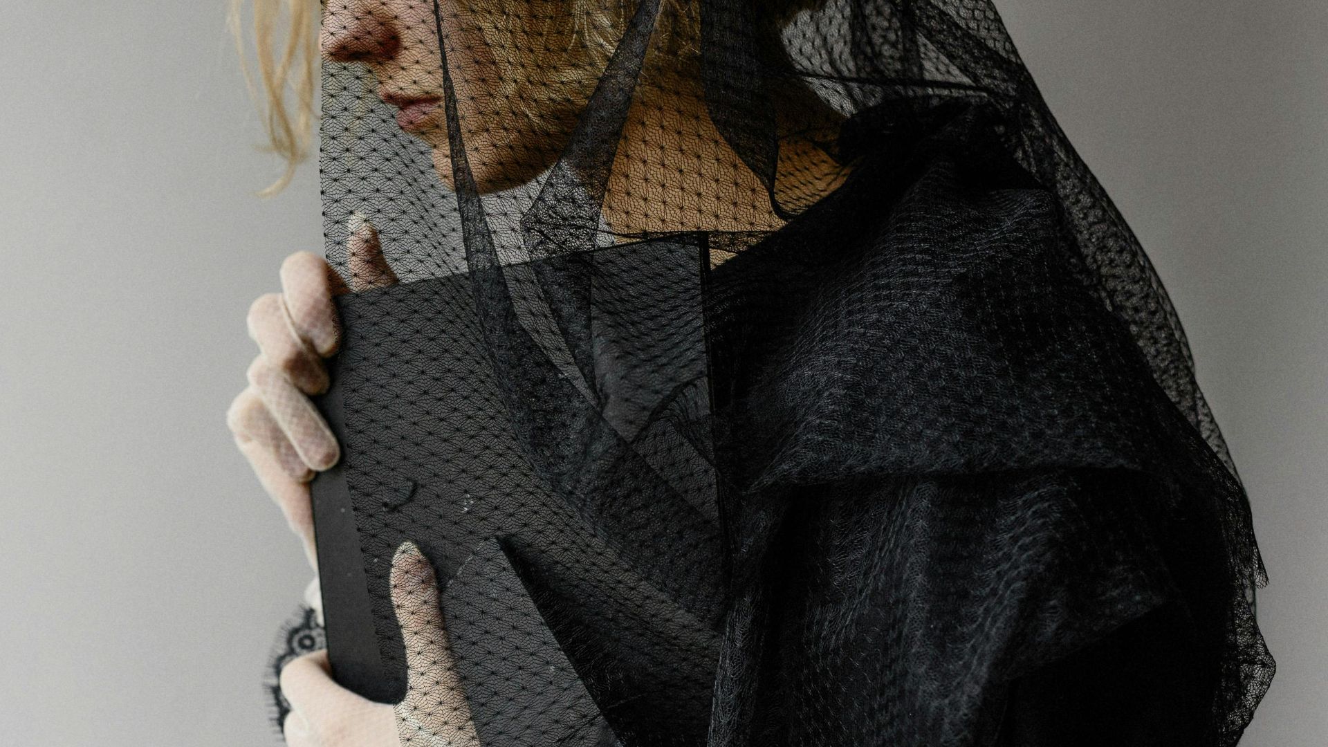 A woman dressed in black mourning attire holds a picture frame beside white flowers.
