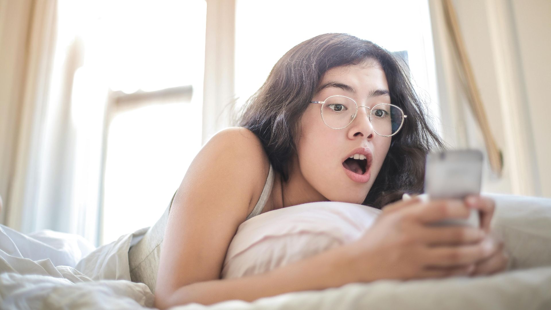 Young woman lying on bed, surprised by something on her smartphone, in a bright bedroom setting.