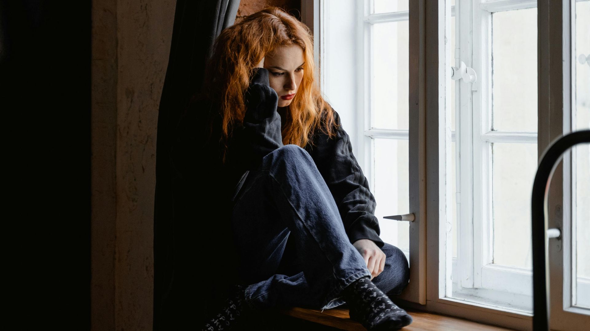 A woman sits by a window, lost in deep thoughts and moody light.
