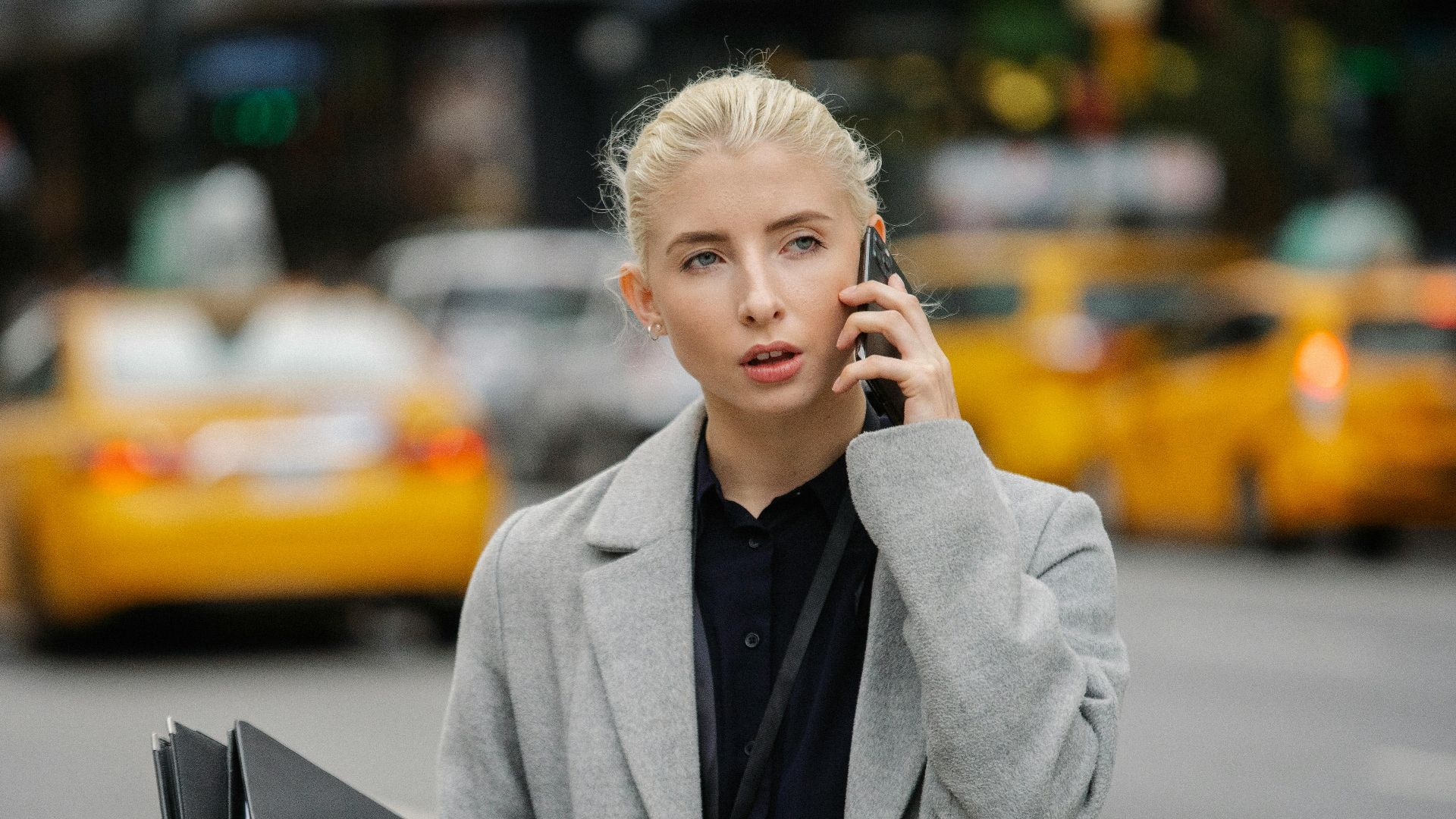 Focused young businesswoman in gray coat standing on busy street with folders and having conversation on mobile phone
