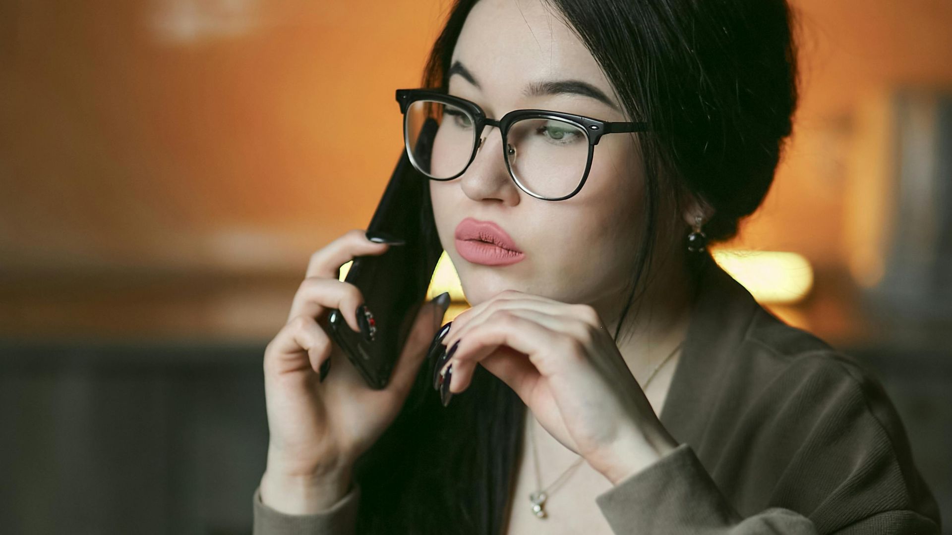 A woman with eyeglasses engaged in a phone call indoors, focused and thoughtful.