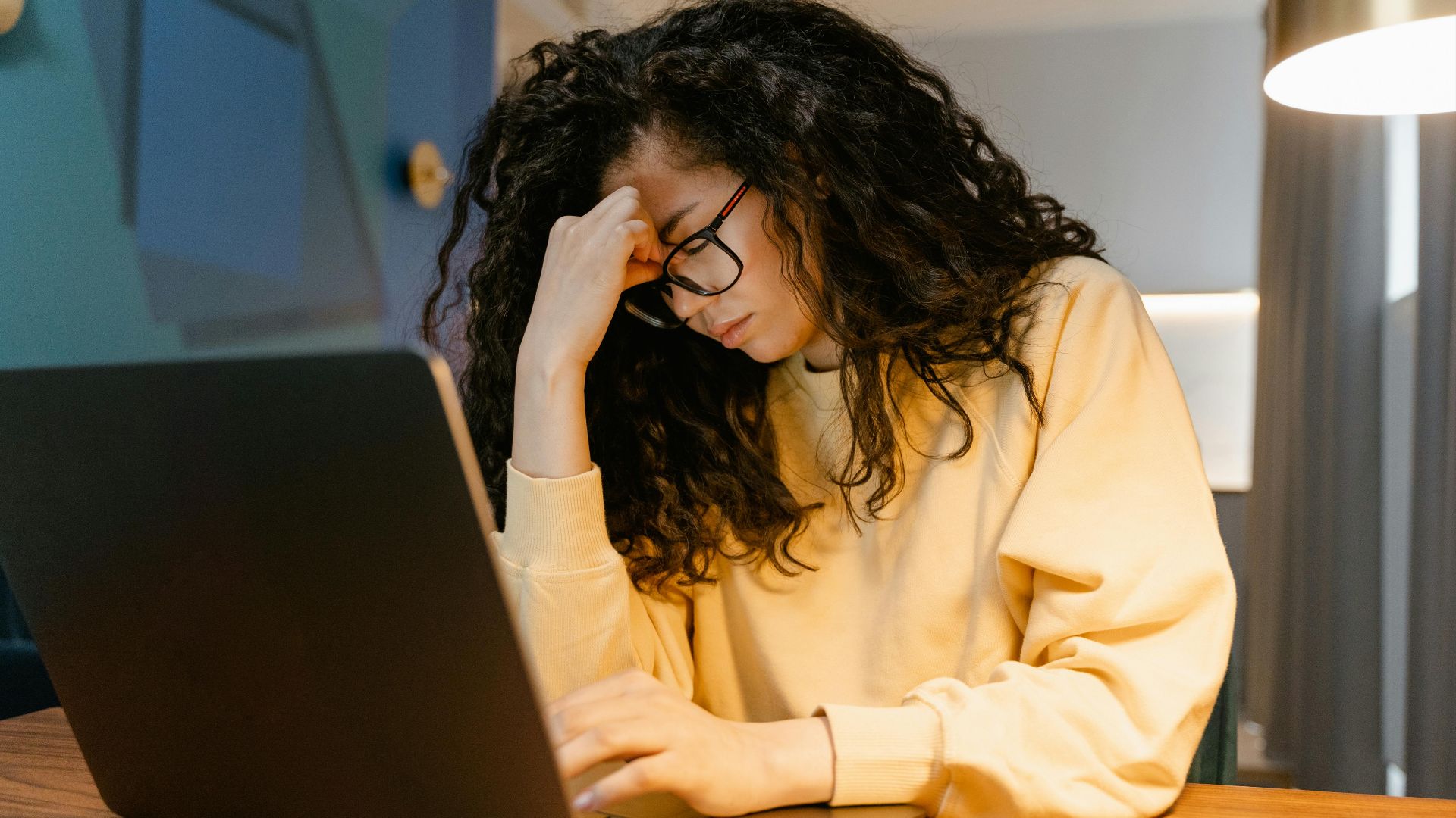 A tired woman with curly hair and glasses experiencing stress while working on a laptop.