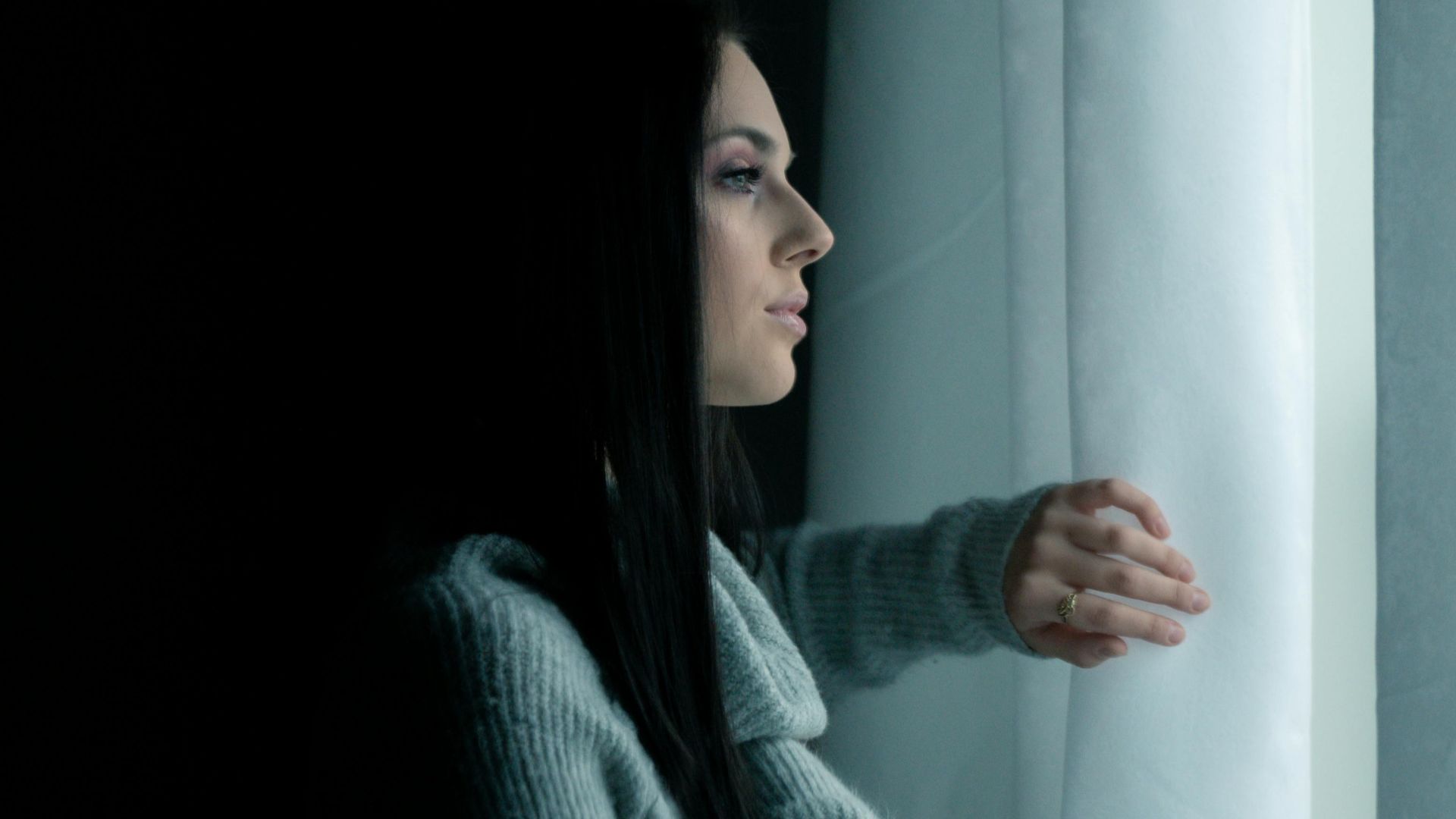Portrait of a woman looking out a window with a thoughtful expression in a dimly lit room.