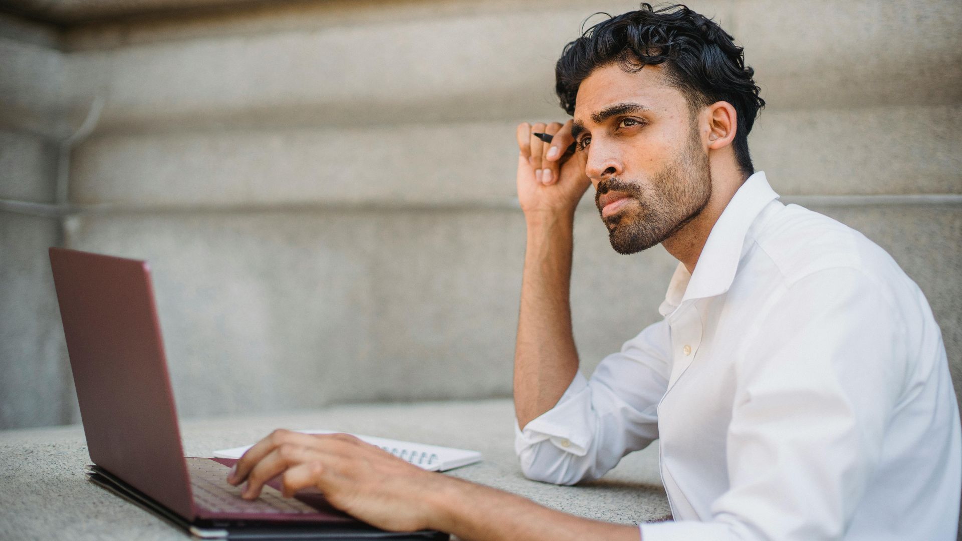 Professional man in a white shirt working on a laptop, contemplating ideas in an urban setting.