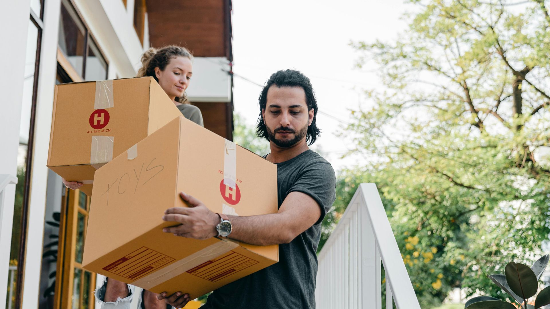 Young multiracial boyfriend and girlfriend in casual wear carrying big carton boxes and watching step on porch stairs while moving out of old home on sunny day in summer