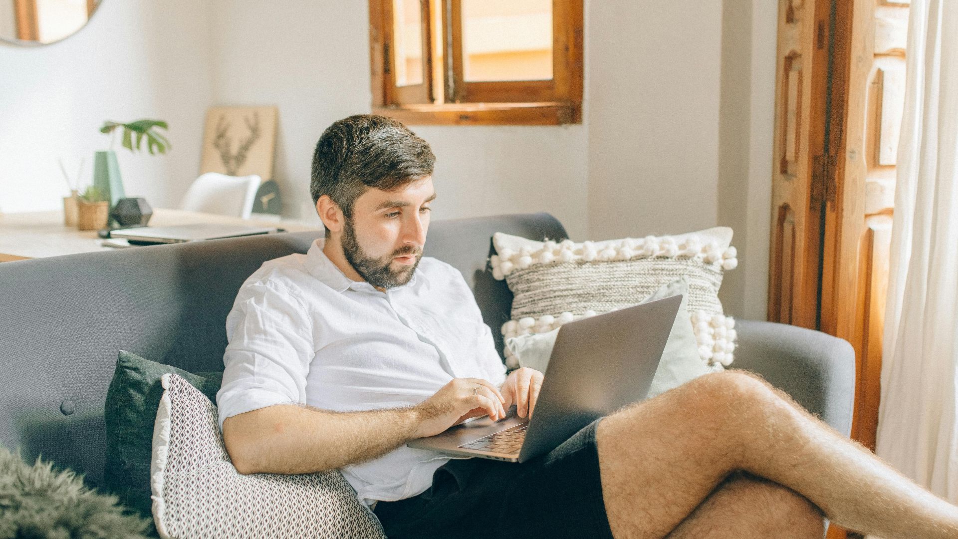 Adult man sitting on couch using laptop, working remotely from cozy home setting.