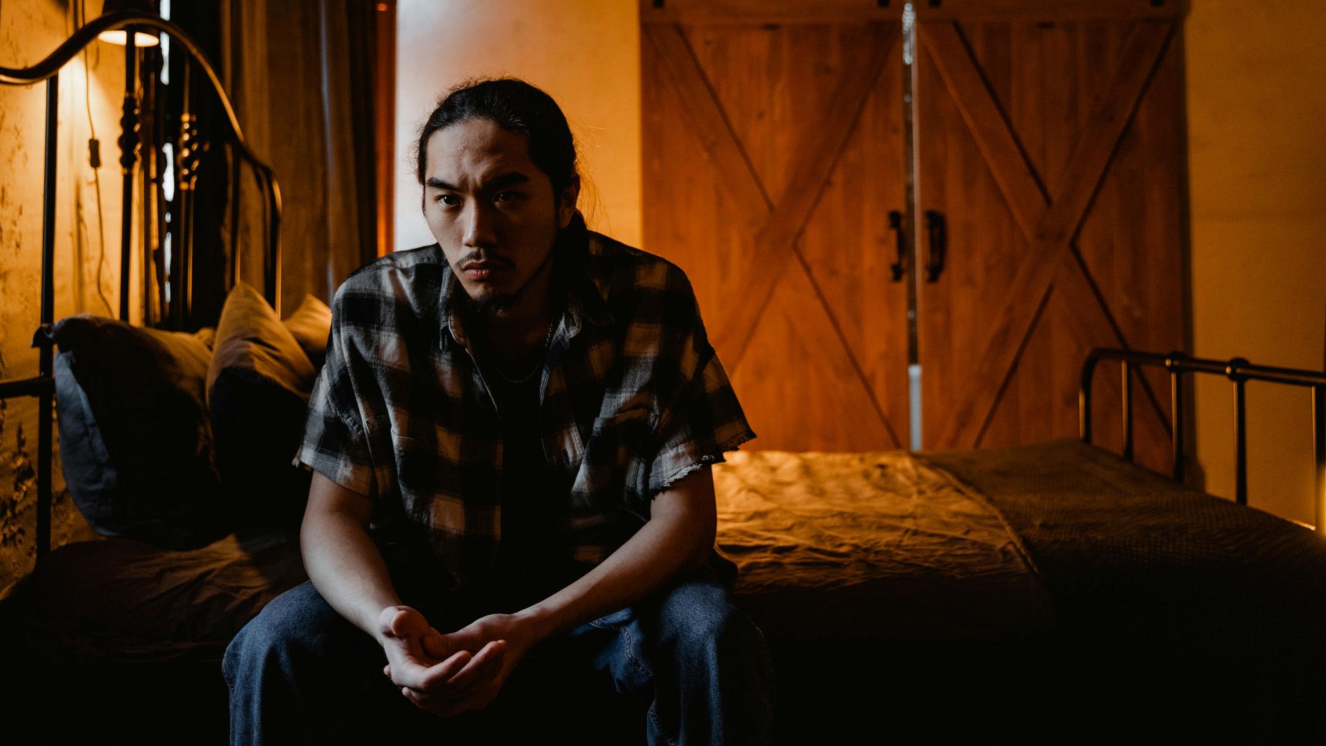 Pensive man sitting on bed in rustic, dimly lit room, creating a reflective mood.