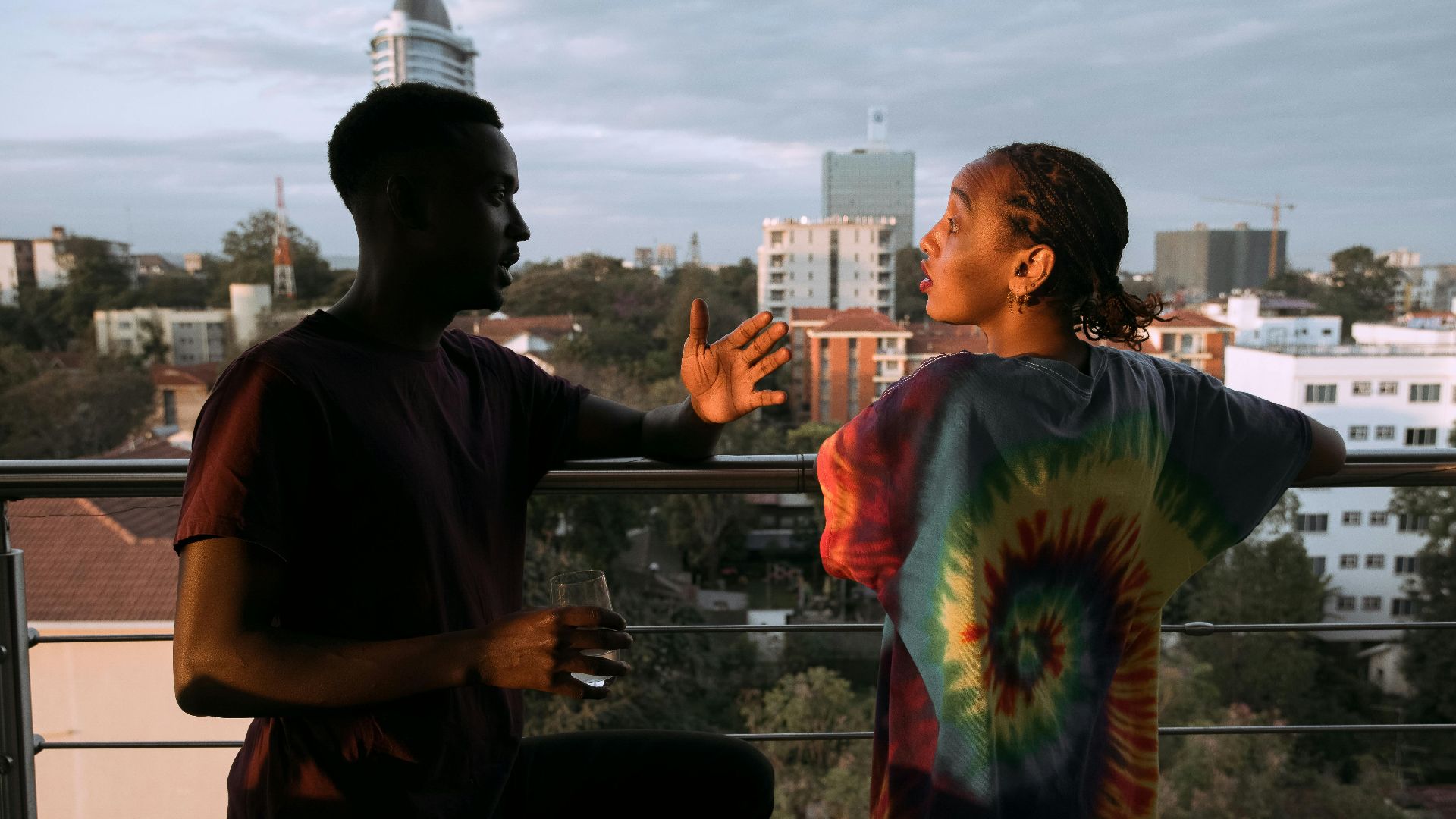 A couple in vibrant attire engaged in conversation on a city balcony at sunset.