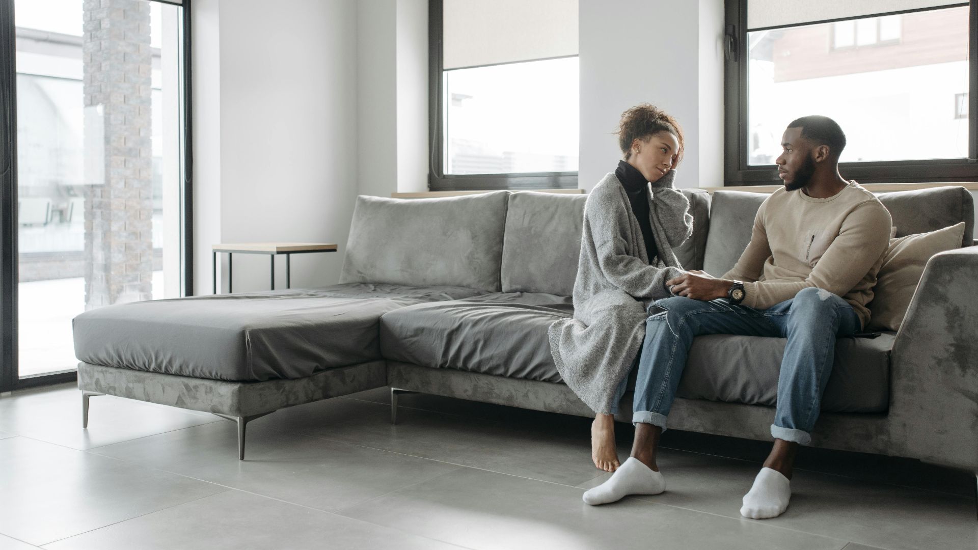 A couple sits together on a contemporary grey sofa in a bright room, sharing a comforting moment.