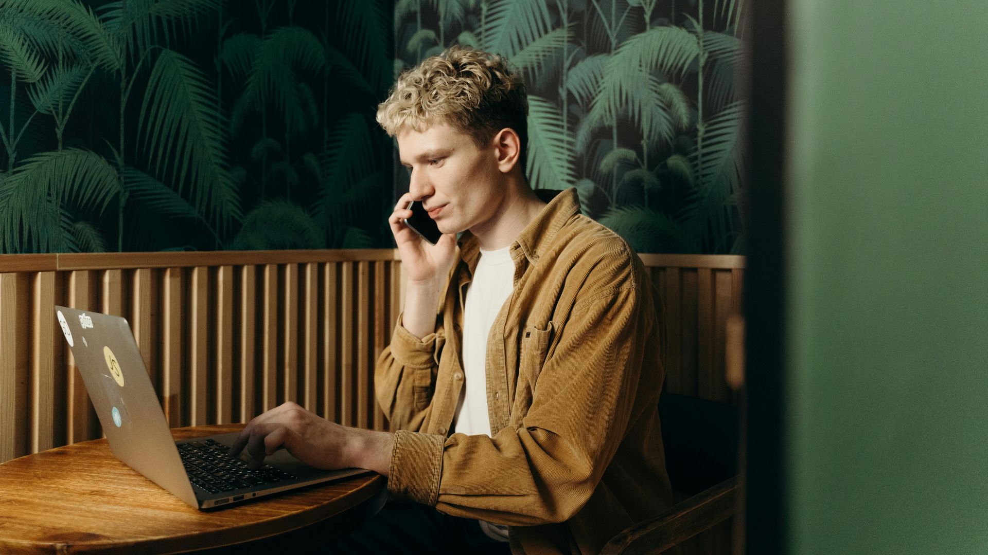 A young man working on a laptop while talking on the phone in a stylish cafe.
