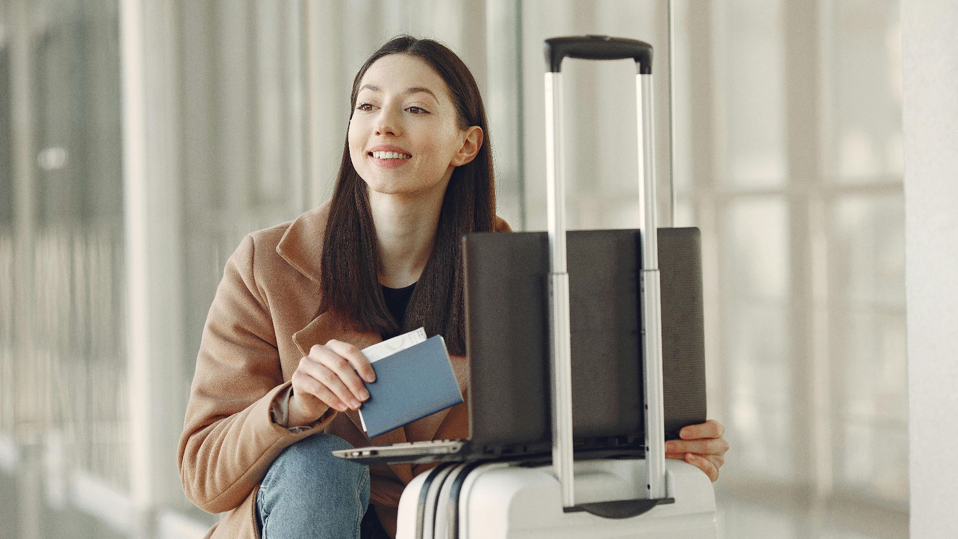 A woman sitting with her laptop and luggage, holding a passport, waiting in an airport terminal.