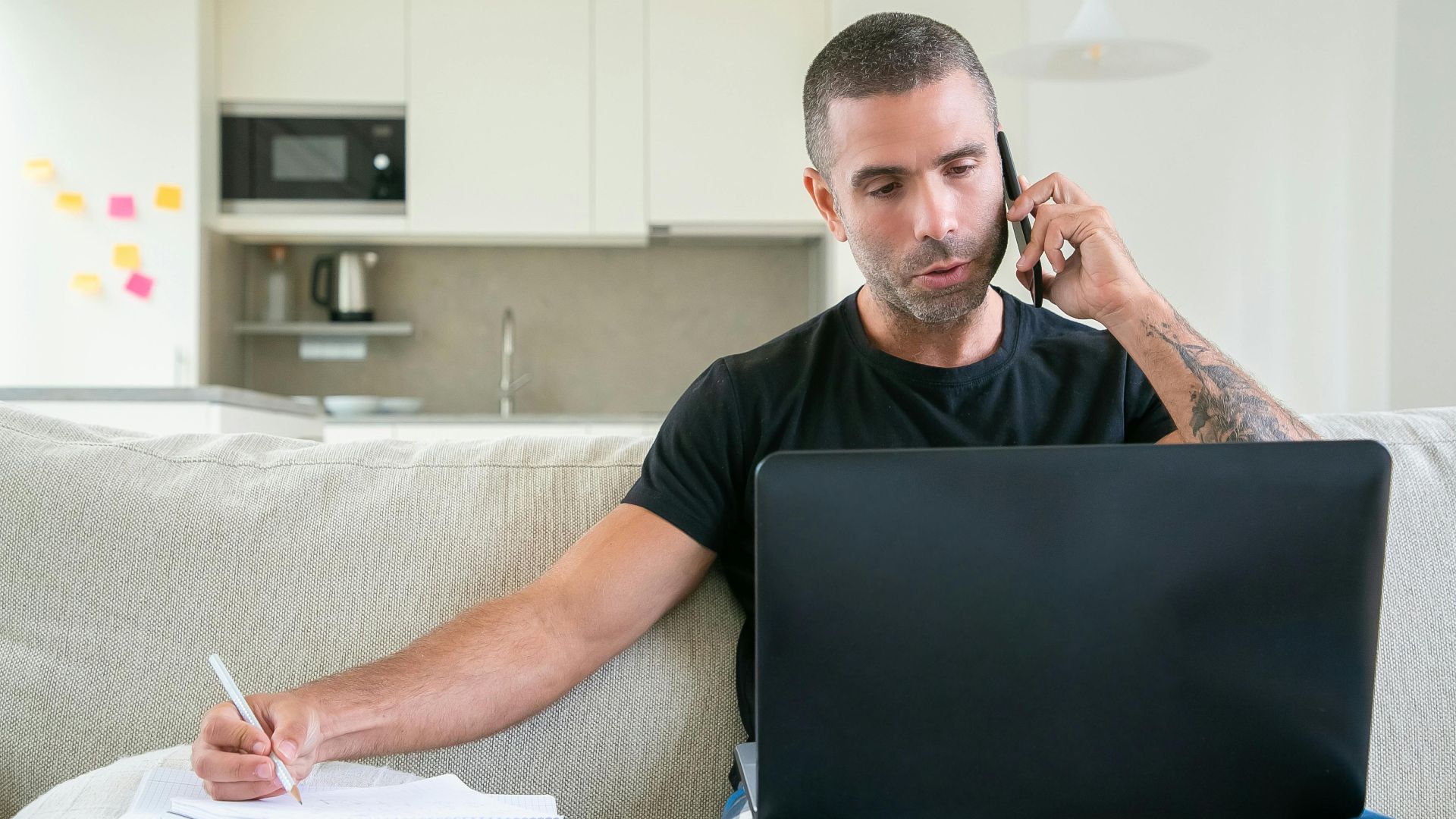 Man working from home using a laptop and smartphone while taking notes in a modern kitchen setting.
