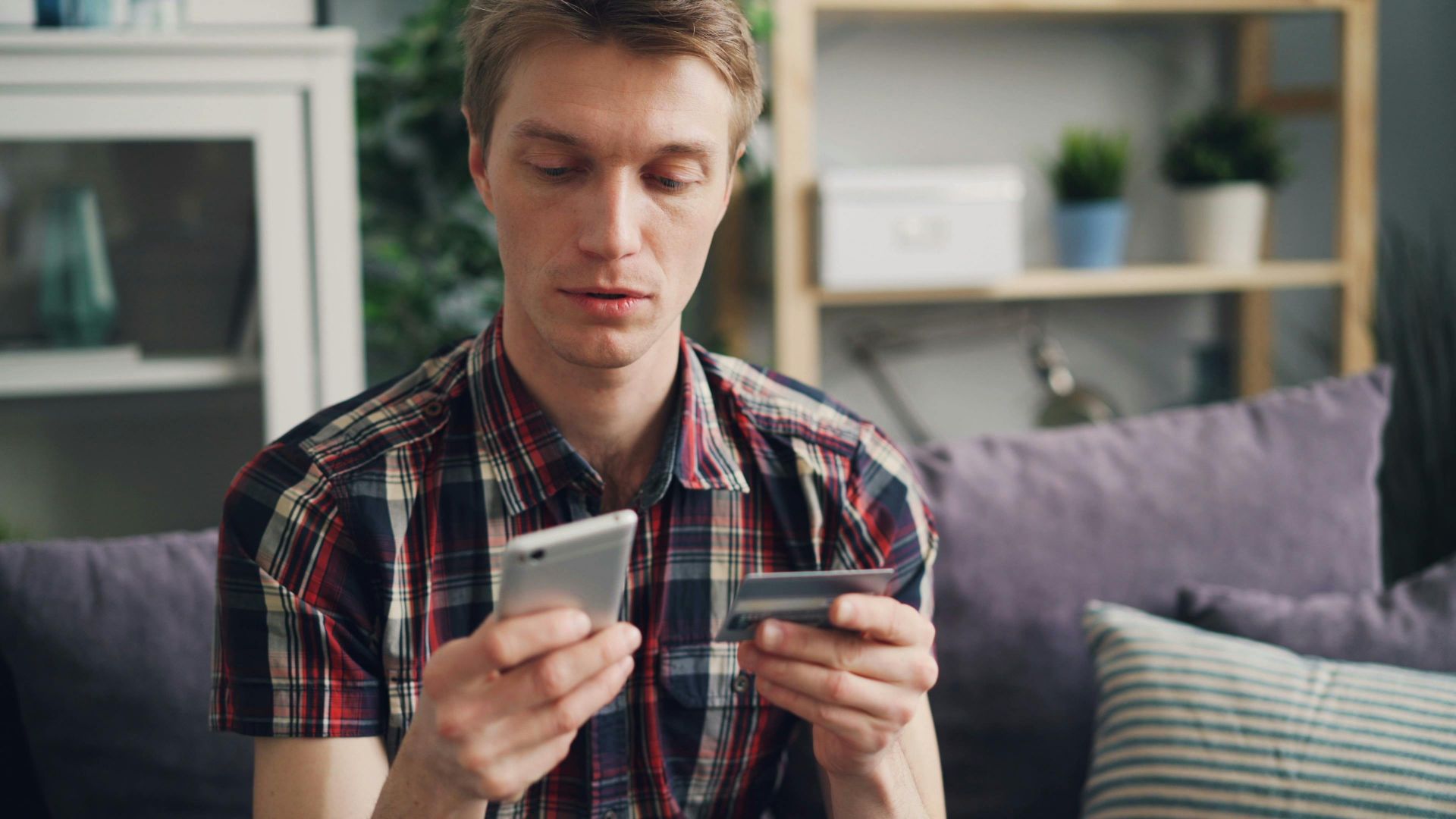 Adult man sitting on sofa using smartphone and credit card for online shopping indoors.