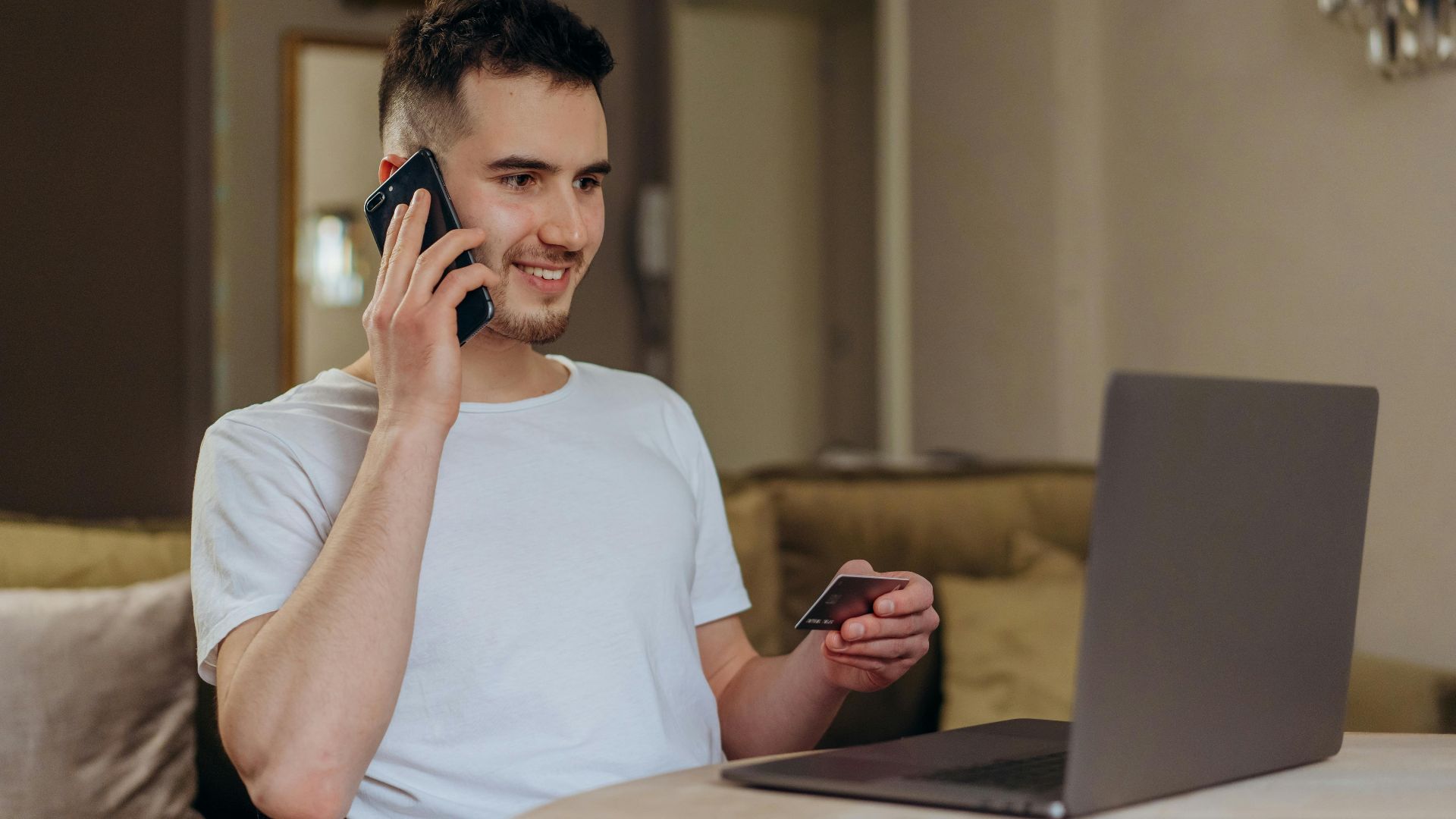 A young man smiles while shopping online using a phone and laptop, holding a credit card.