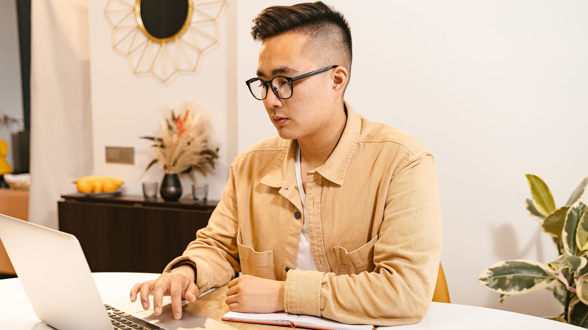 Young Asian man focused on work, using a laptop at a modern home office.