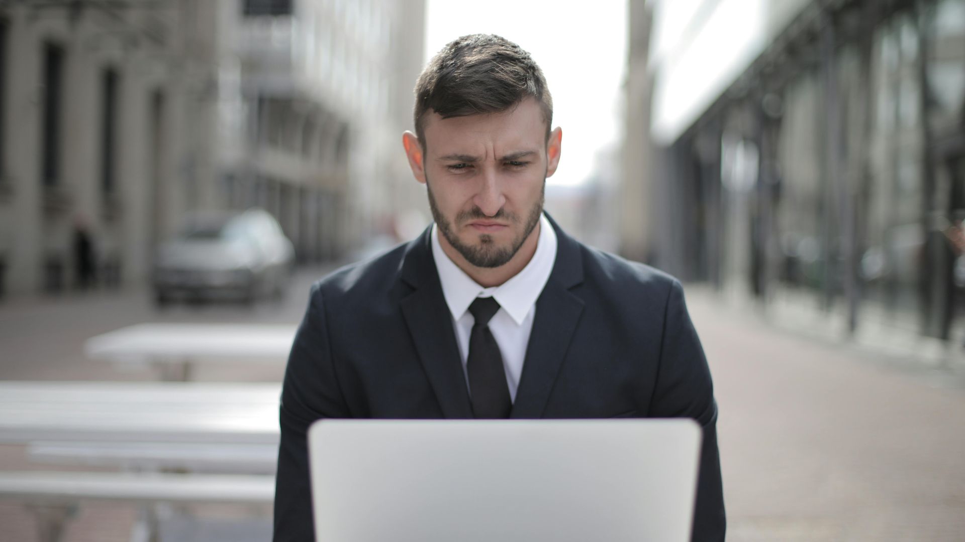 A focused businessman in a suit works on his laptop outdoors in an urban setting.