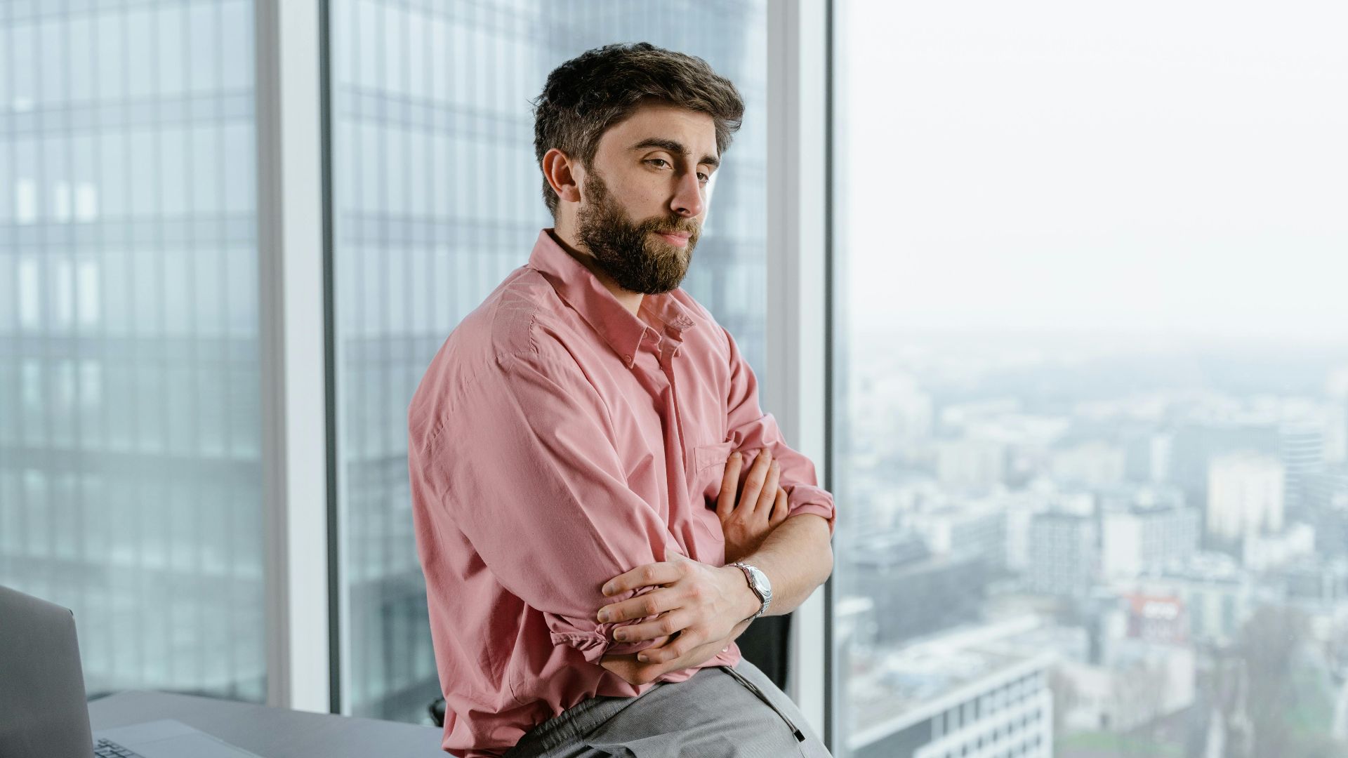 Man in pink shirt sitting on table, arms crossed, overlooking city from high-rise office.