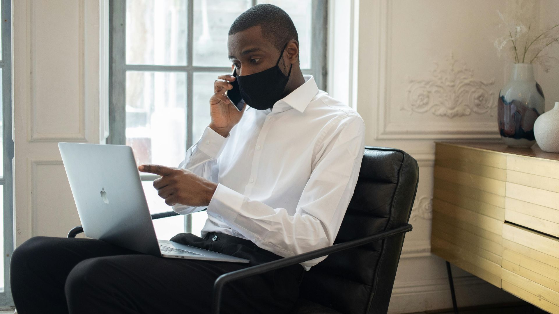 Businessman in mask talking on phone while using laptop indoors. Modern remote work setup.