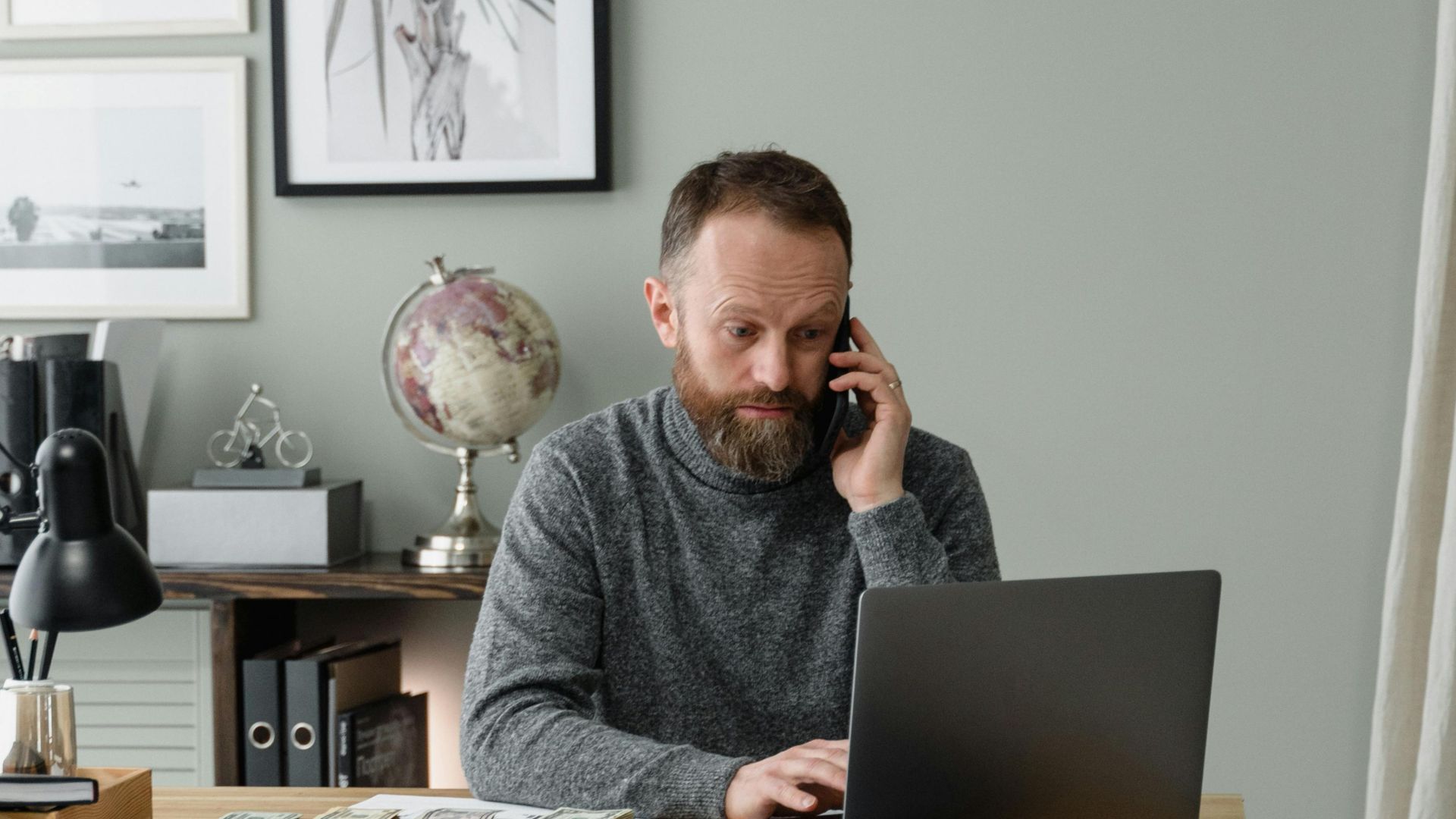 A man working in a modern office, multitasking with a phone and laptop at a wooden desk.