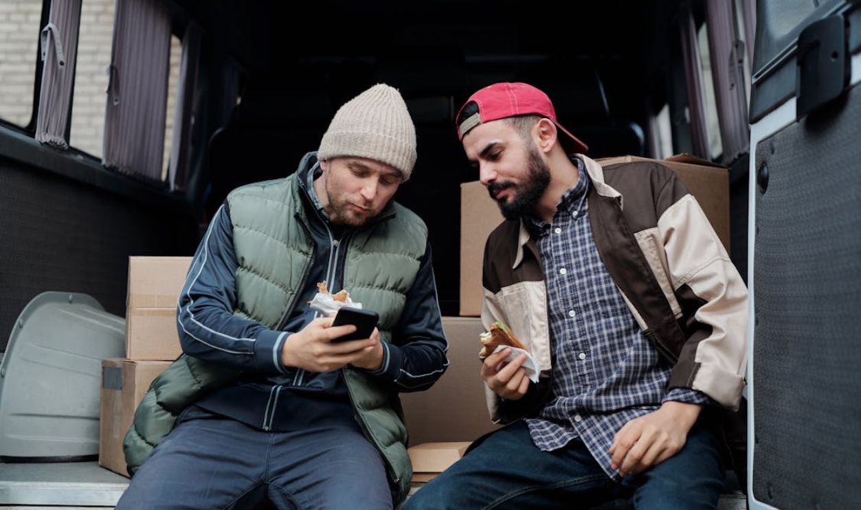 Men Sitting on the Back of a Van Eating Sandwiches and Looking at a Smartphone Screen
