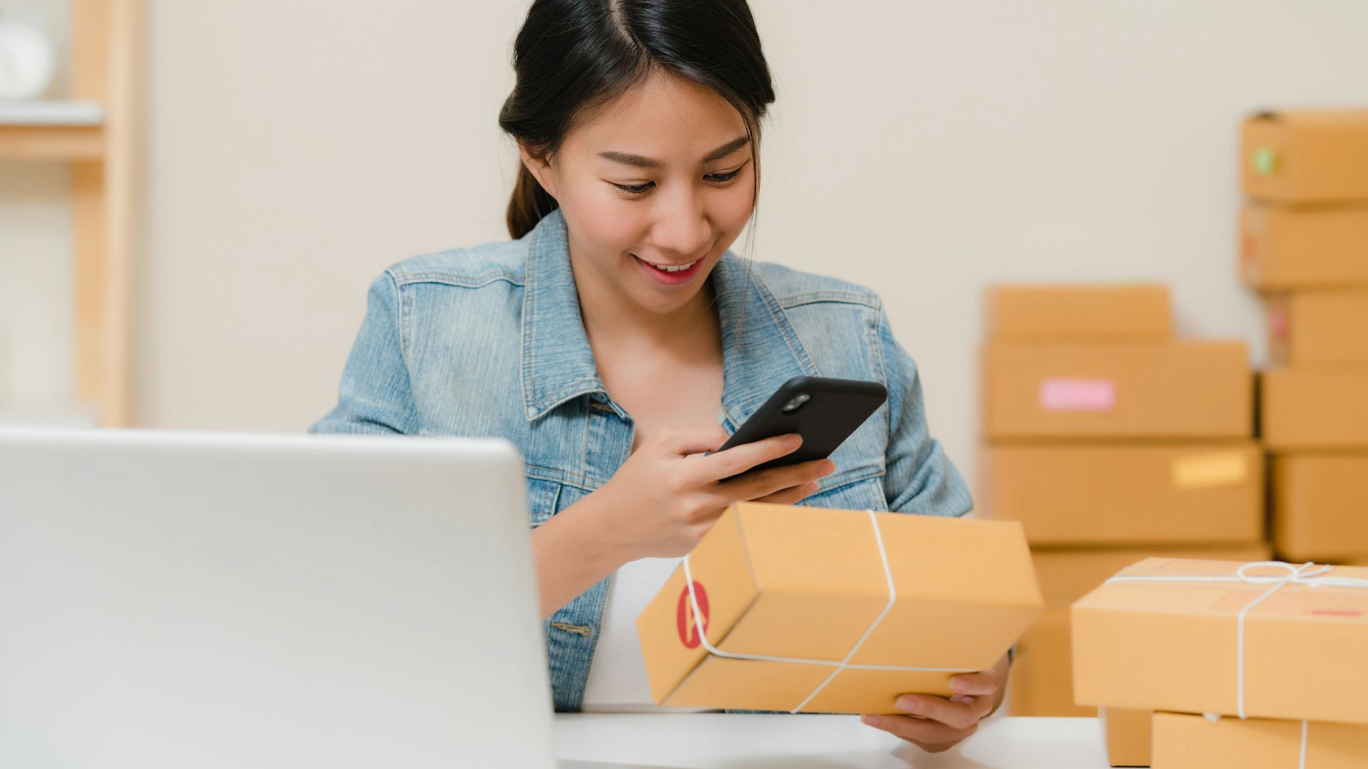Woman checking package with phone near laptop and boxes