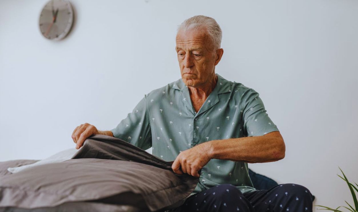 An Elderly Man in Gray Shirt and Black Pants Sitting on the Bed