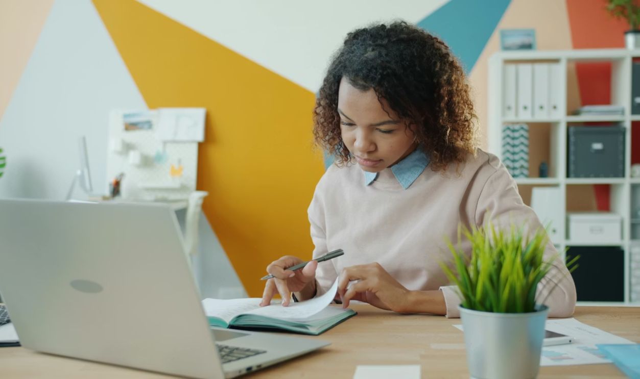 Serious young Afro-American woman is writing in notebook working in office alone concentrated on business information. People and workplace concept.