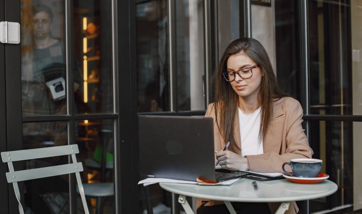 Woman Wearing Eyeglasses Sitting in the Coffee Shop Using Her Laptop and Taking Notes