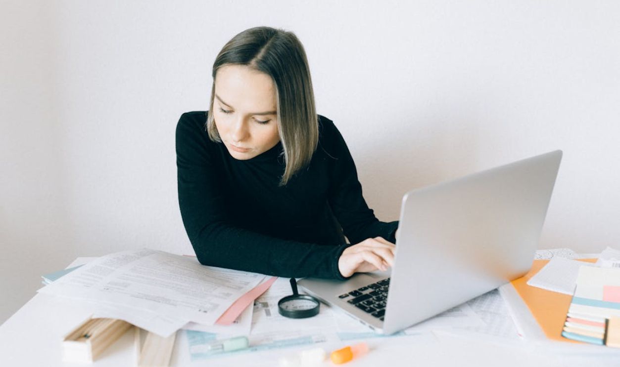 A Woman in Black Long Sleeves Doing Paperwork while Typing on Her Laptop