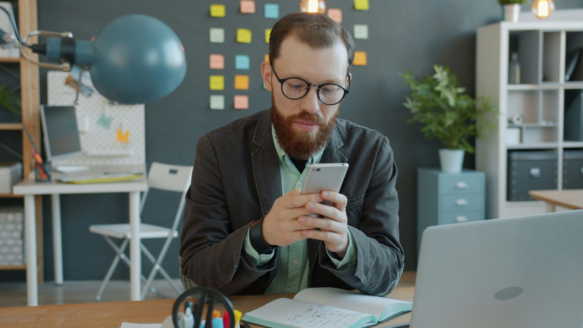 Man in glasses using smartphone at office desk.
