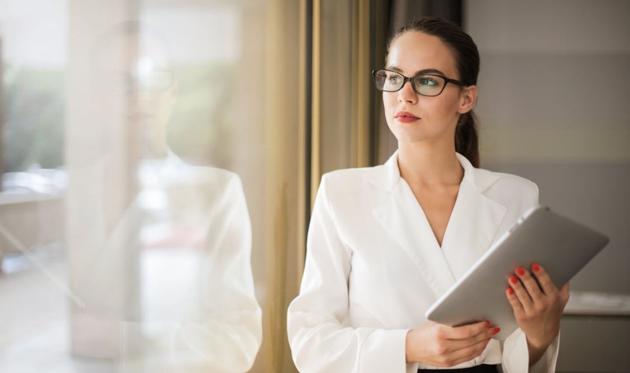 Photo of Woman in White Top and Glasses Holding a Tablet While Looking Outside a Window