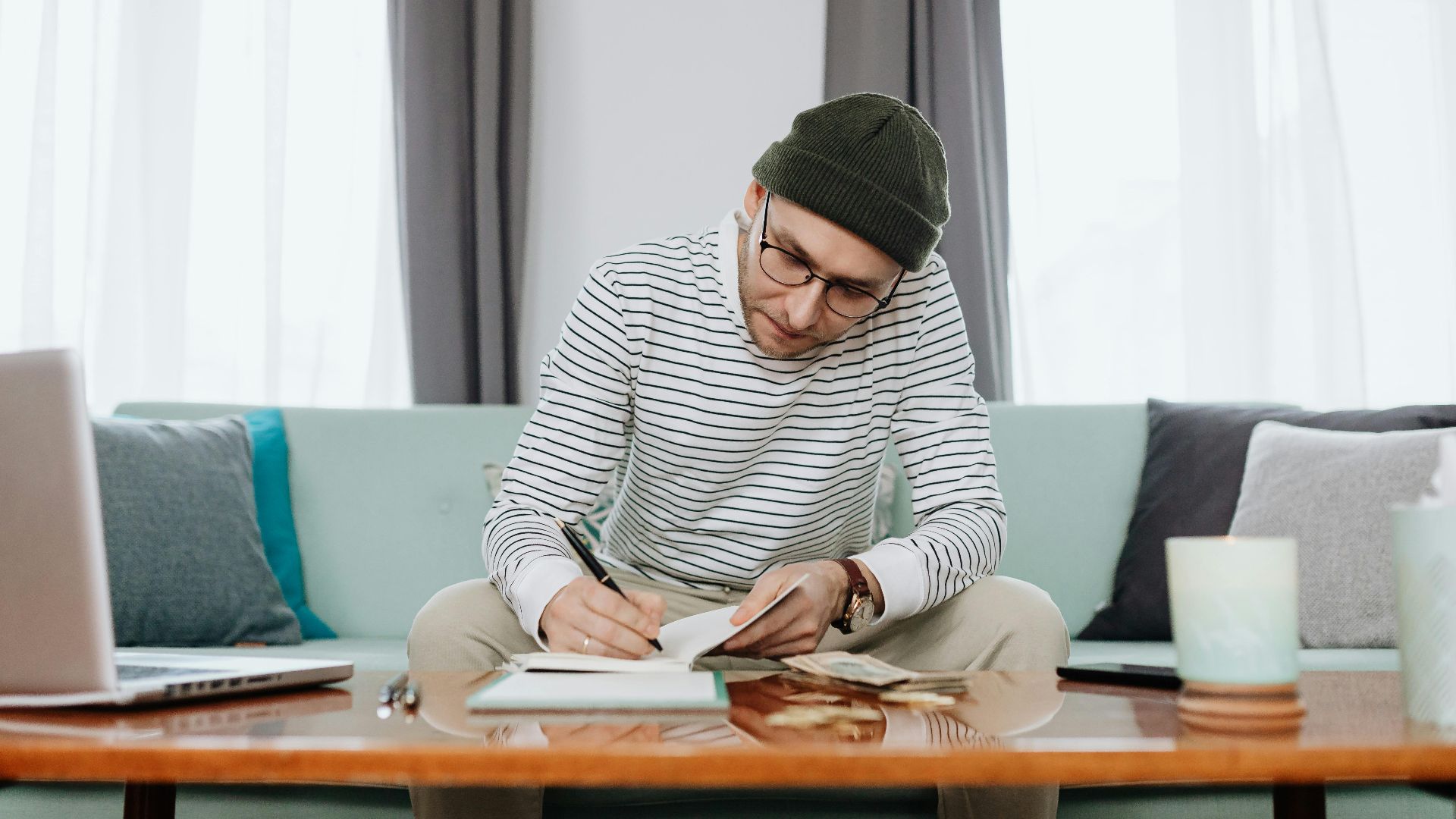 A man in a beanie writing notes and budgeting at a living room desk with a laptop.