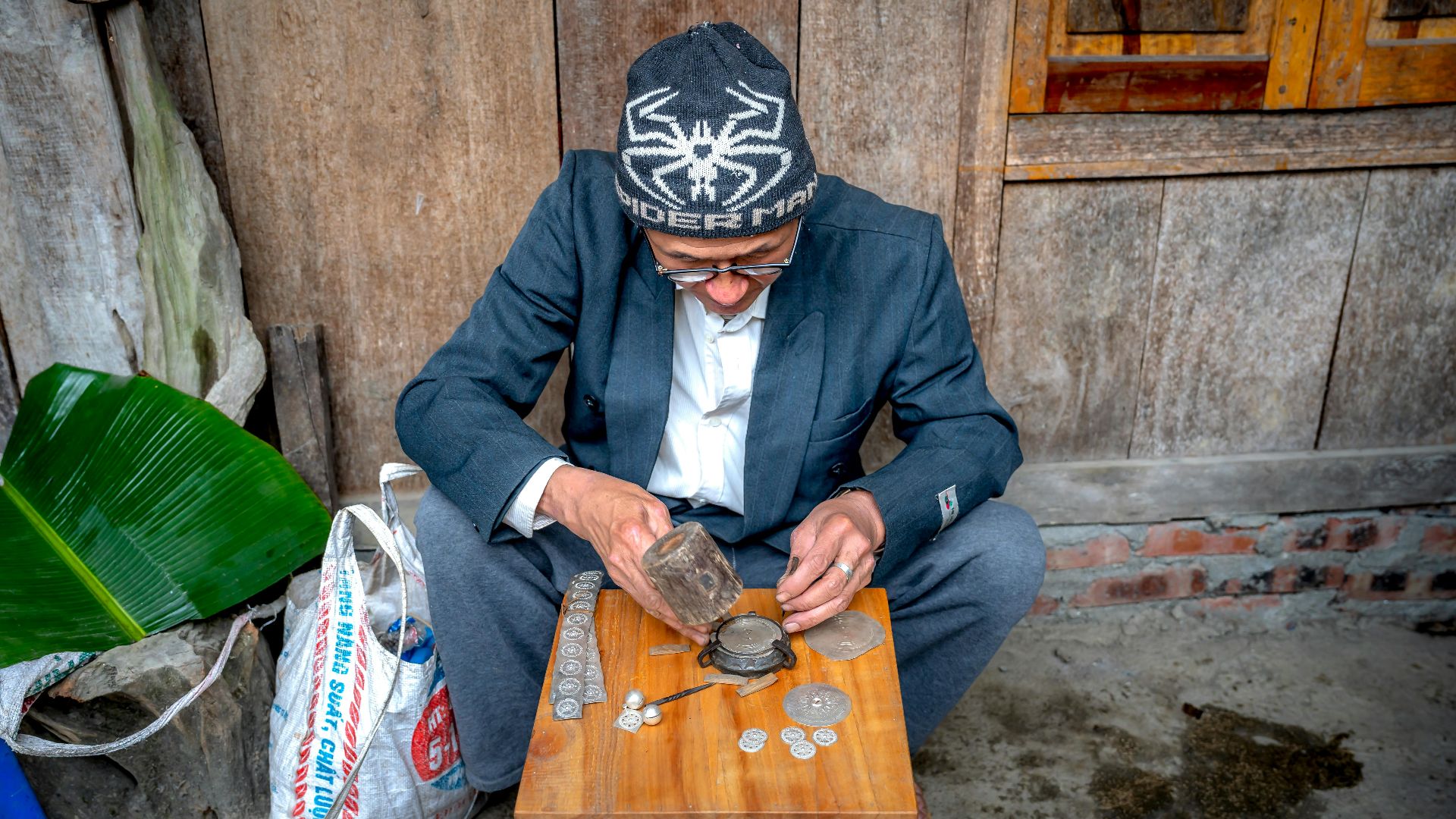Senior man in winter cap working on metal items with coins on a wooden table in a rustic setting, focused on craftsmanship.