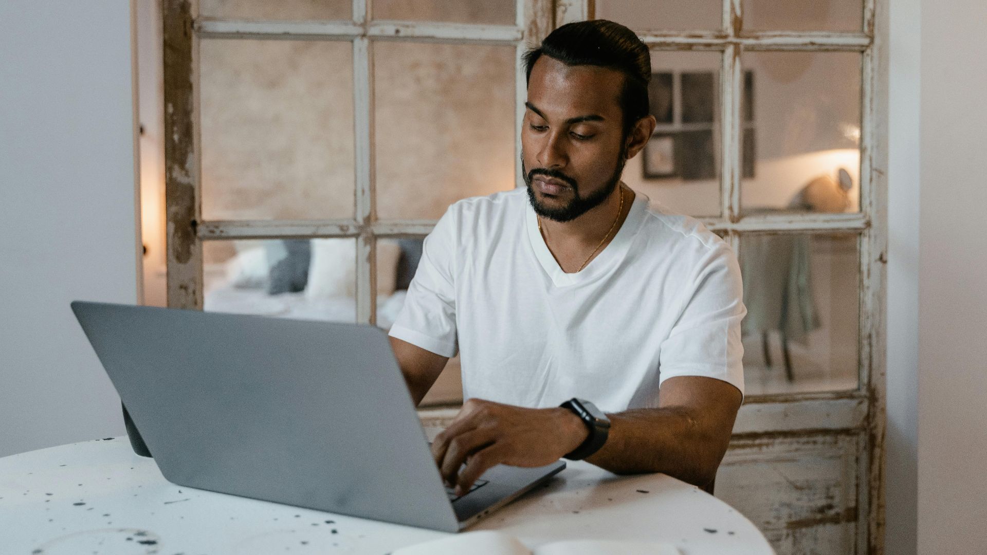 Adult man with beard focused on laptop work at home desk during the day.