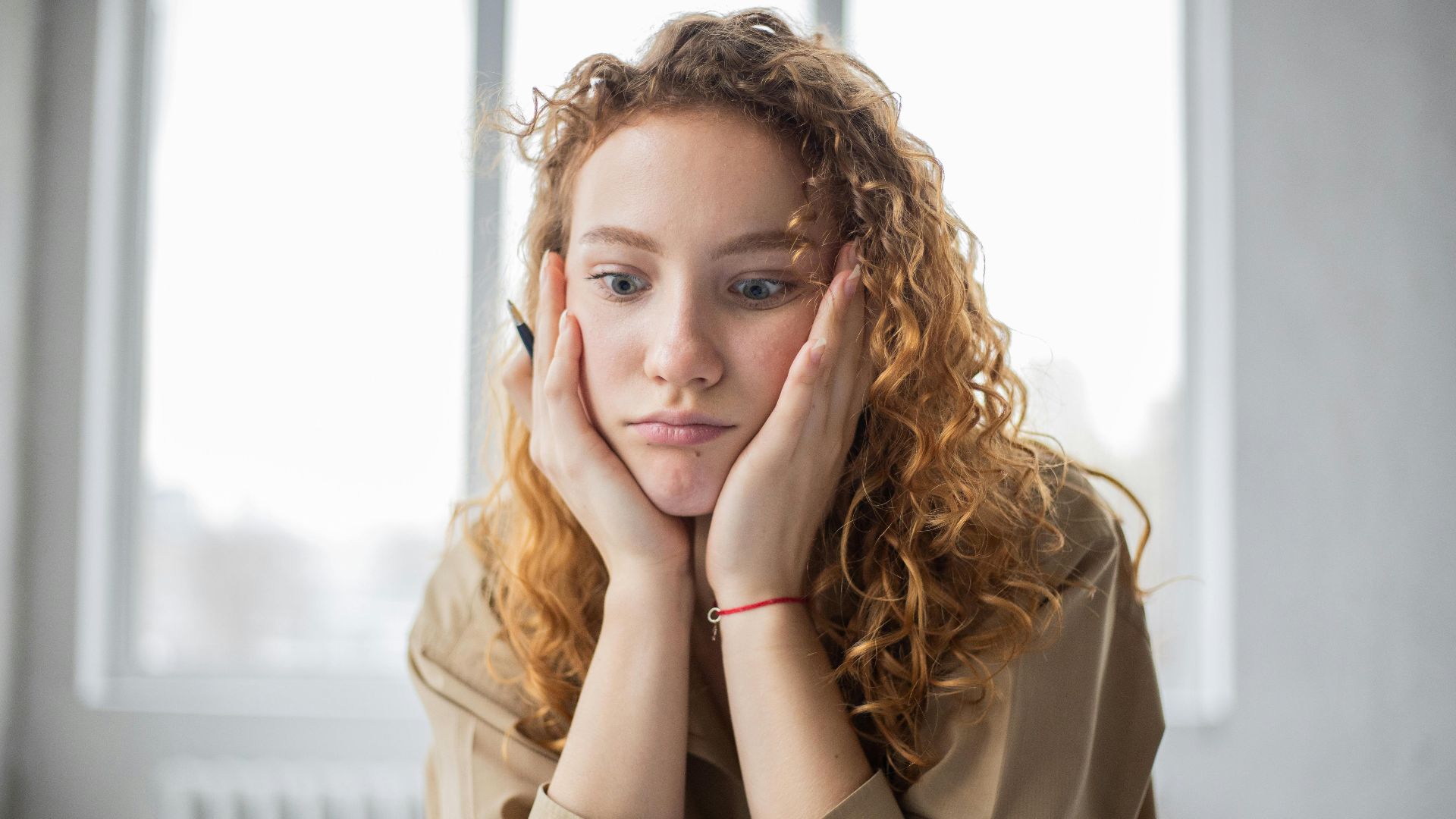 Pensive female student with hands on cheeks looking down while sitting in light room on blurred background while deciding task in frustration