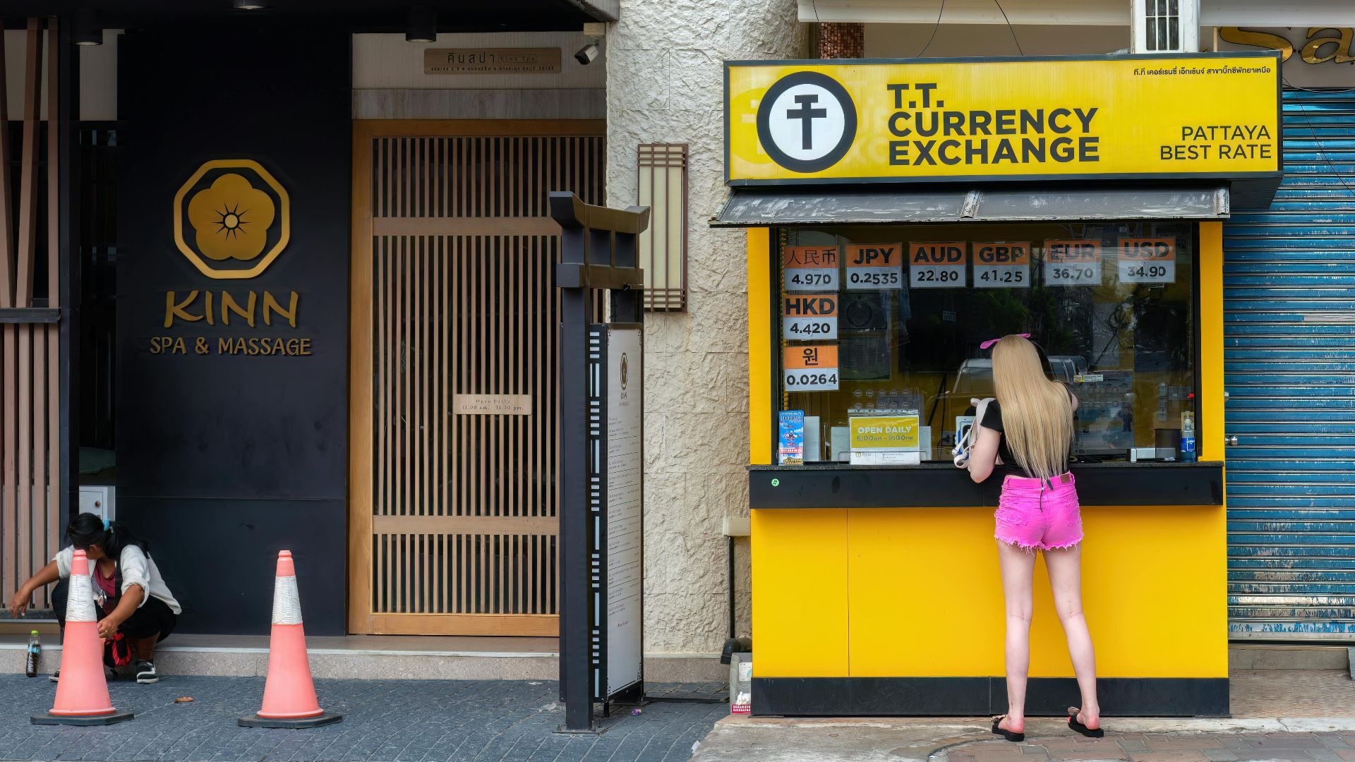 Woman at currency exchange booth in Pattaya, Thailand, with currency rates displayed.