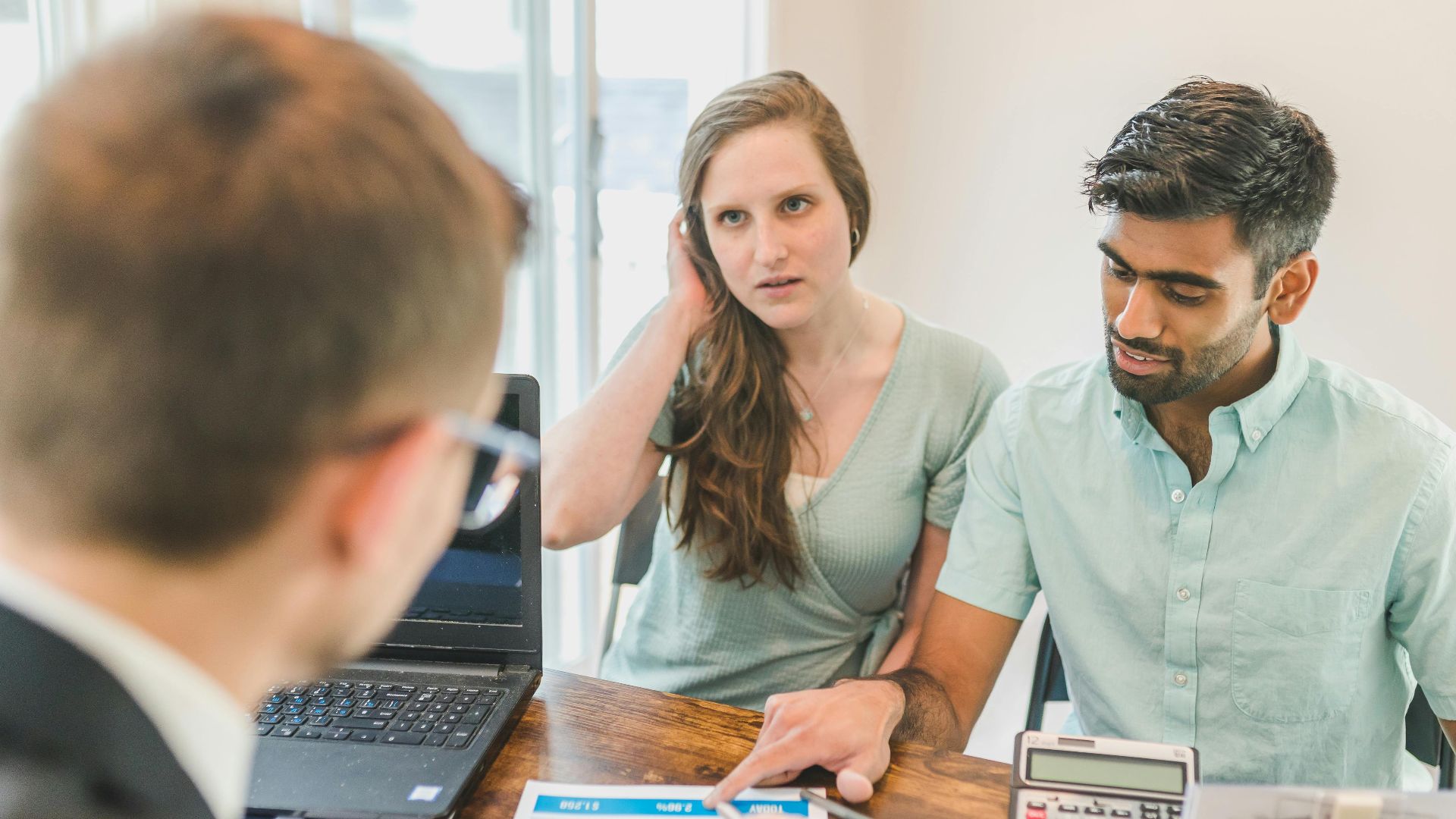 A young couple consults with a realtor about buying a home, indoors in a modern office setting.