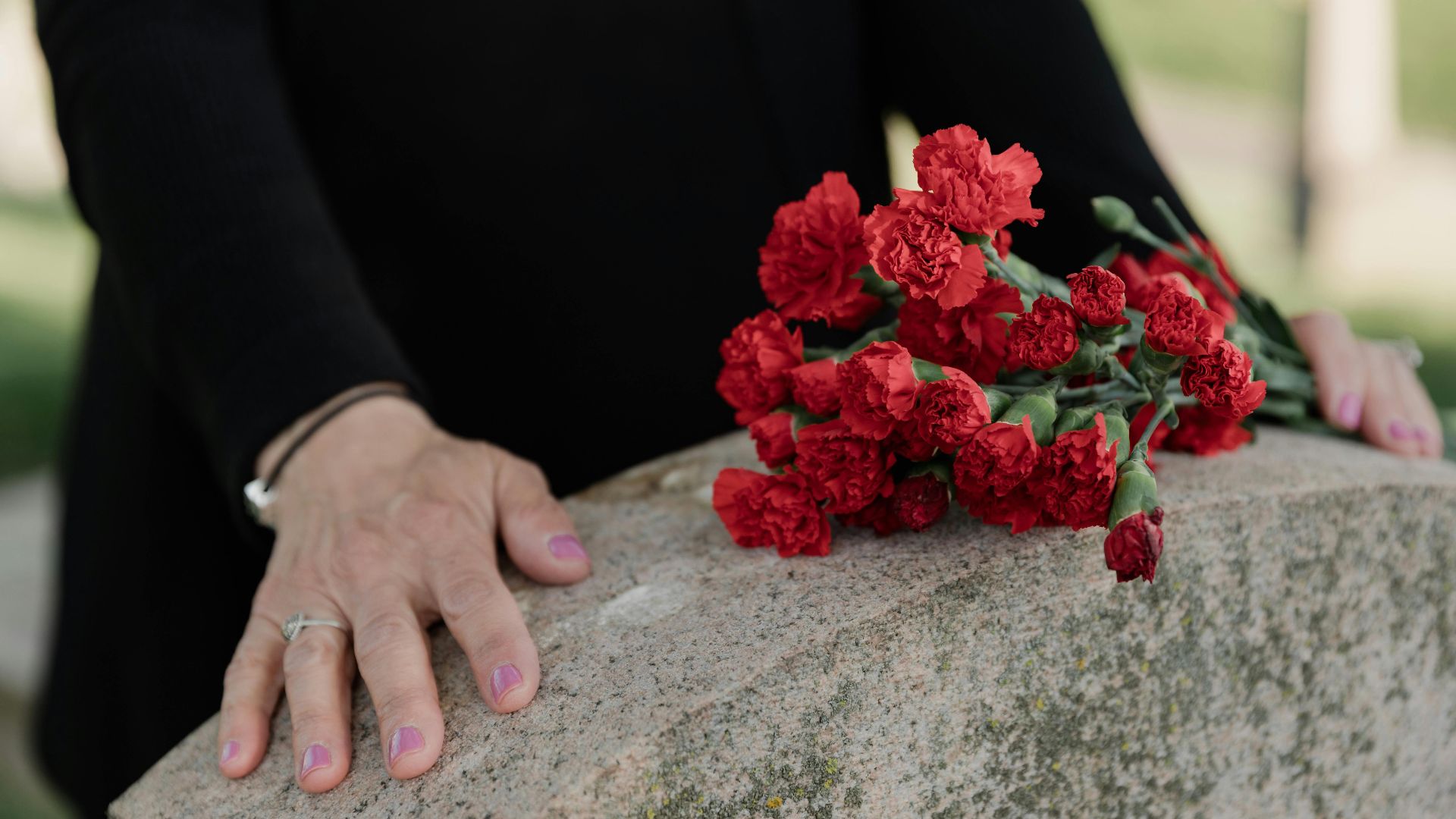 A woman dressed in black stands solemnly by a tombstone with red carnations.