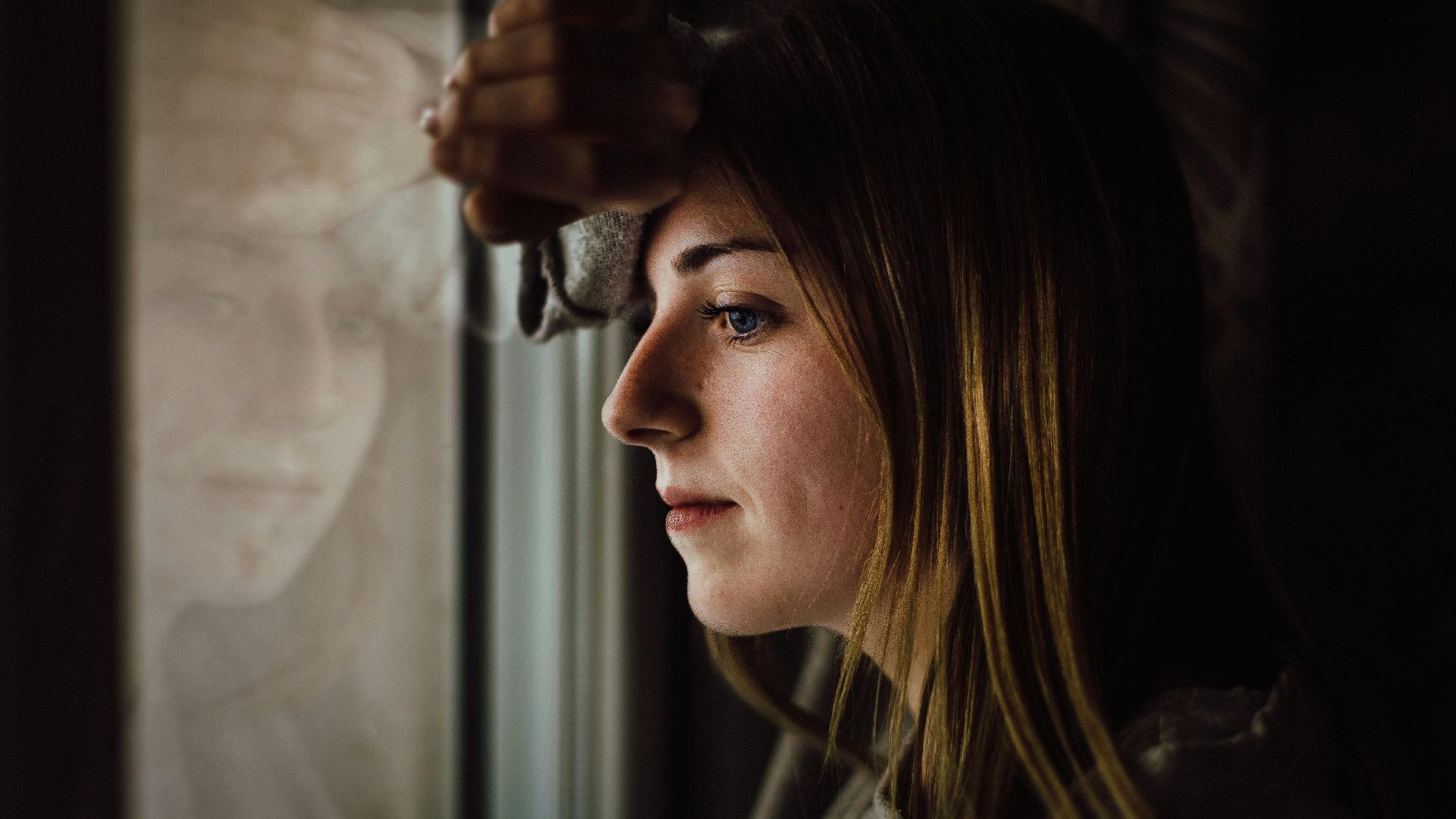 A contemplative young woman looks out a window with a reflective expression, indoors.
