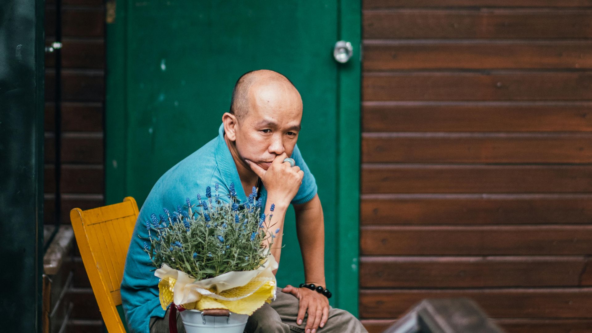Bald man in casual shirt seated outdoors by a wooden cabin, contemplating.