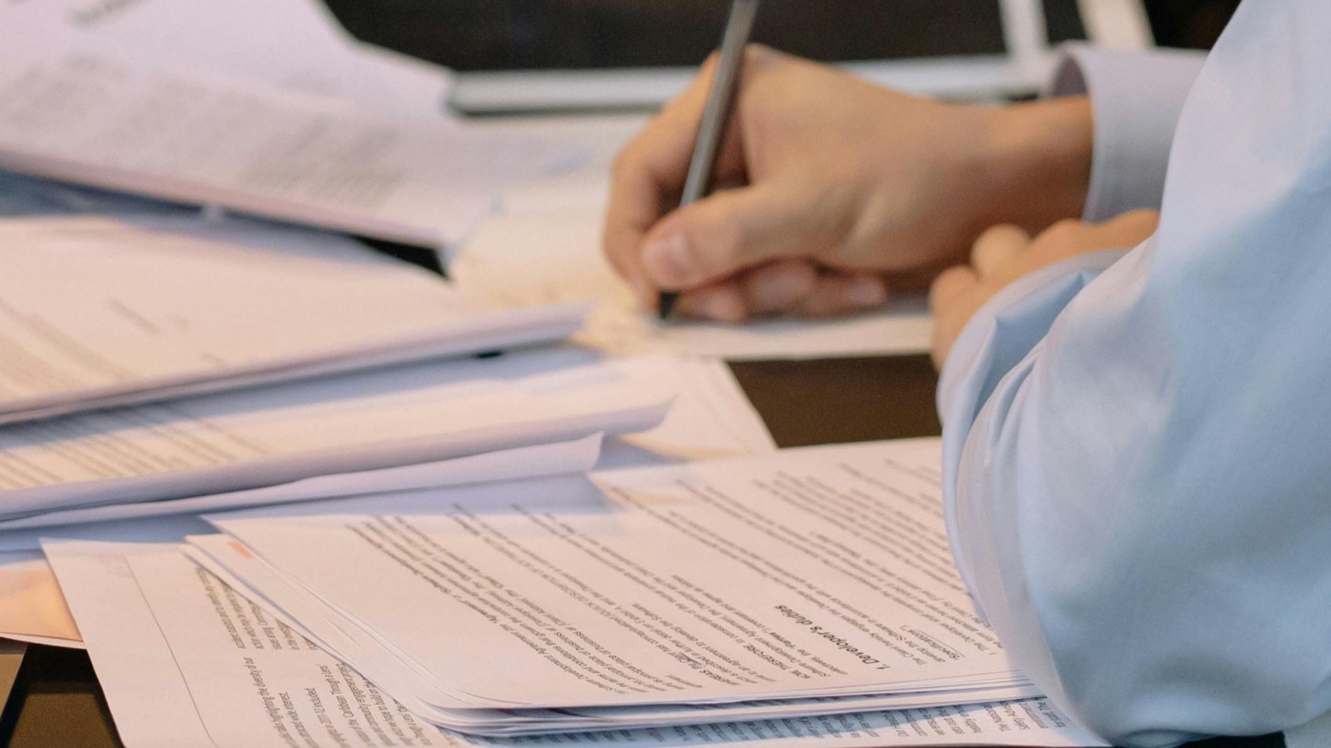 A young professional takes notes at a desk filled with documents in an office.