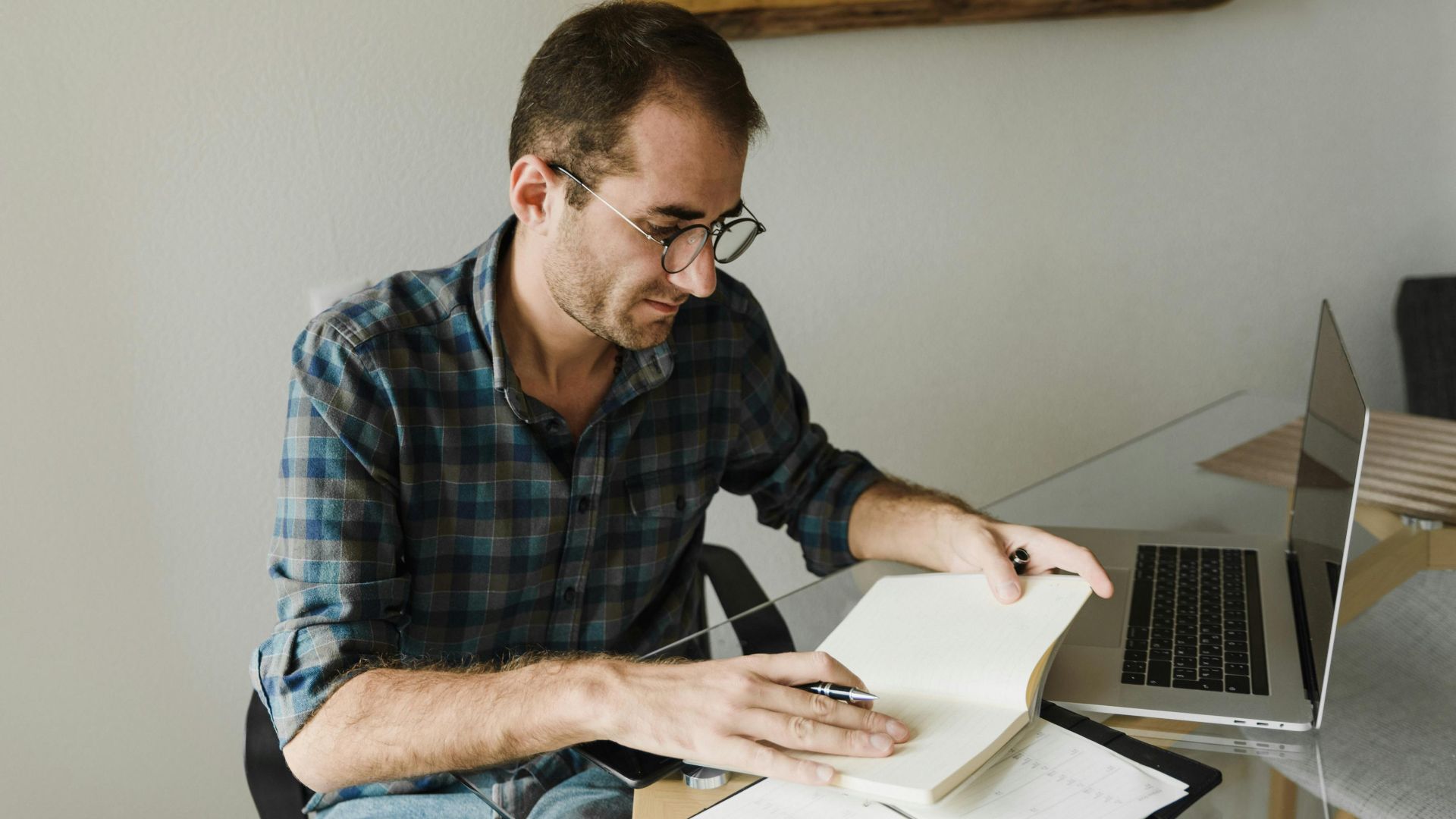 Caucasian man in eyeglasses working on a notebook and laptop in a home office setting.