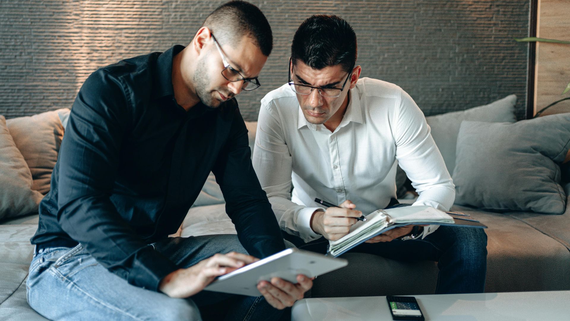 Two men in a modern setting collaborate using a tablet and notebook, emphasizing teamwork and technology.