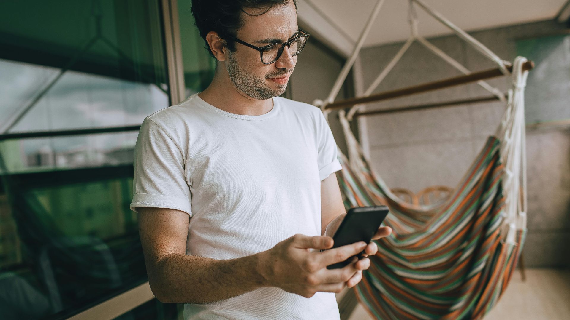 Portrait of a young man with glasses using smartphone near a colorful hammock on a balcony.