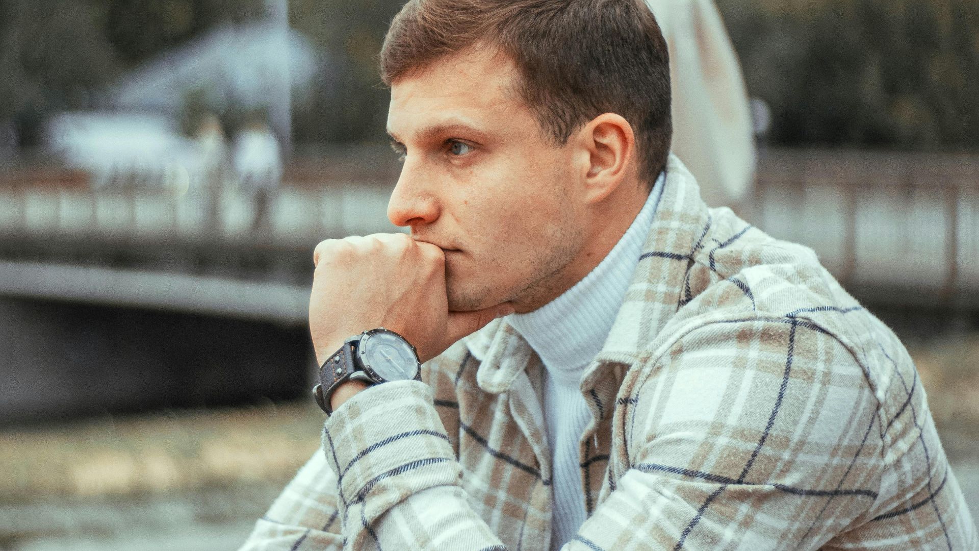 Close-up of a man in a plaid jacket, deep in thought at an outdoor cafe in a city.